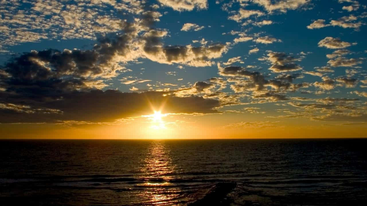 Sunset View 桌布 壁紙 — 海洋上的日落，金色光線反射在水面和深藍色天空中散佈的雲朵上。