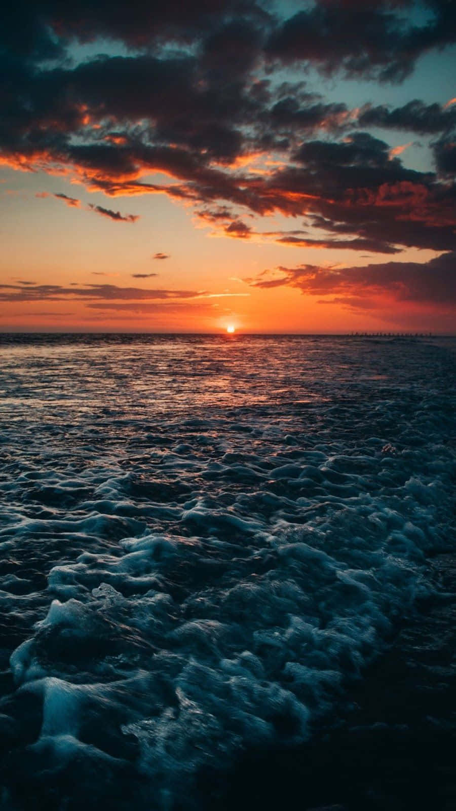海灘 桌布 壁紙 — 海洋上空戲劇性的日落，帶有火熱的橙色雲朵和深藍色的波浪拍打向海岸。