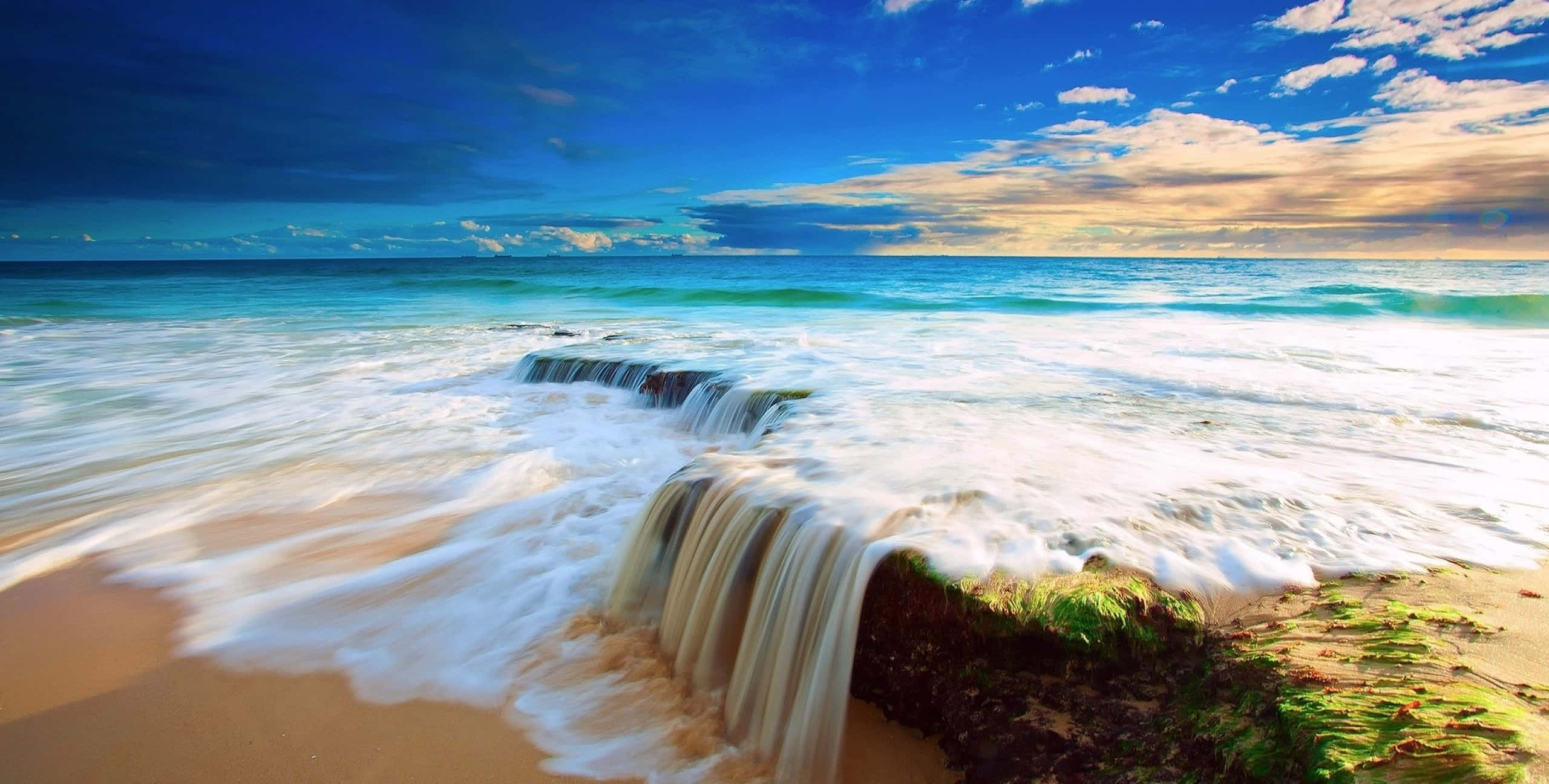 Beautiful Sea 桌布 壁紙 — 海浪在岩石上傾瀉而下，落在金色沙灘上，天空湛藍，白雲點綴。