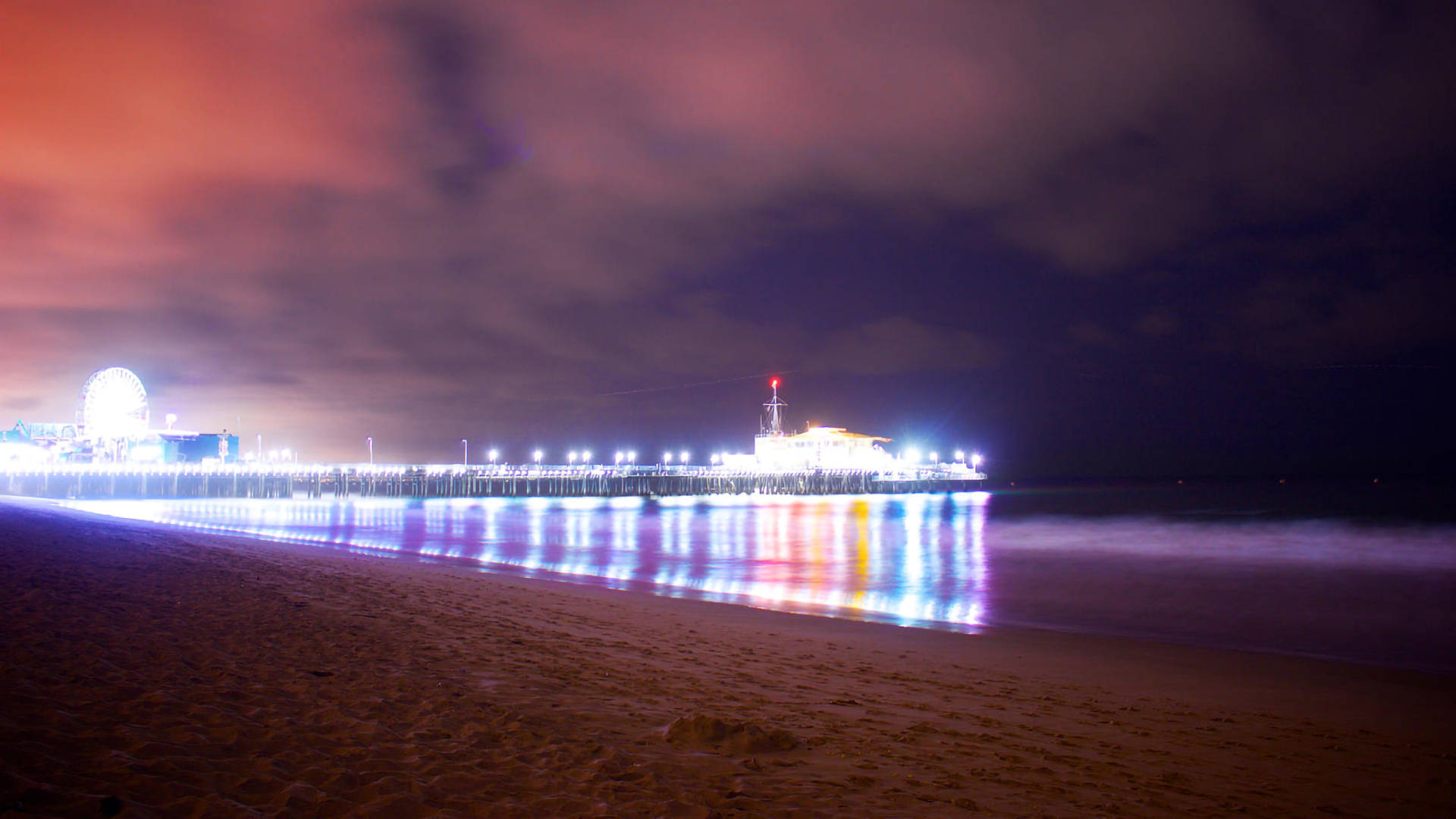 Monica 桌布 壁紙 — 海灘上充滿活力的夜景，明亮的碼頭在色彩繽紛的多雲天空下延伸至海洋。