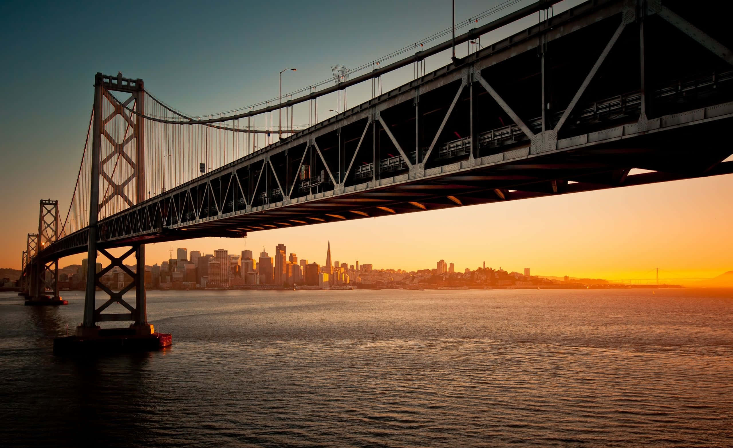 bridge 桌布 壁紙 — 海灣大橋在日落時分橫跨金色水域，背後是舊金山的天際線在溫暖色調中閃耀。