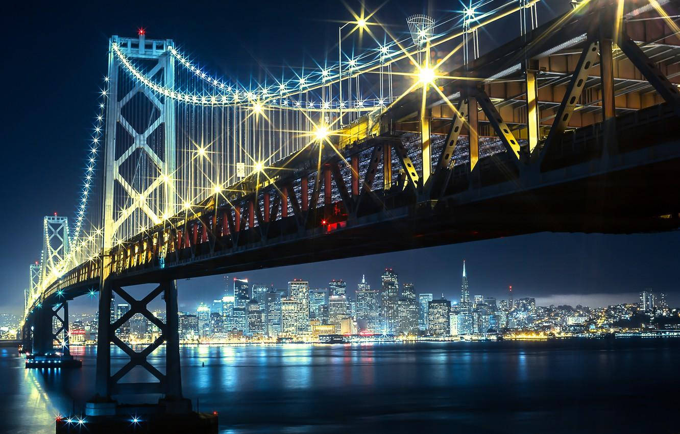 夜空 桌布 壁紙 — 海灣大橋在金色燈光下閃耀的迷人夜景，平靜的水面倒映著燈光，襯托著充滿活力的城市天際線。