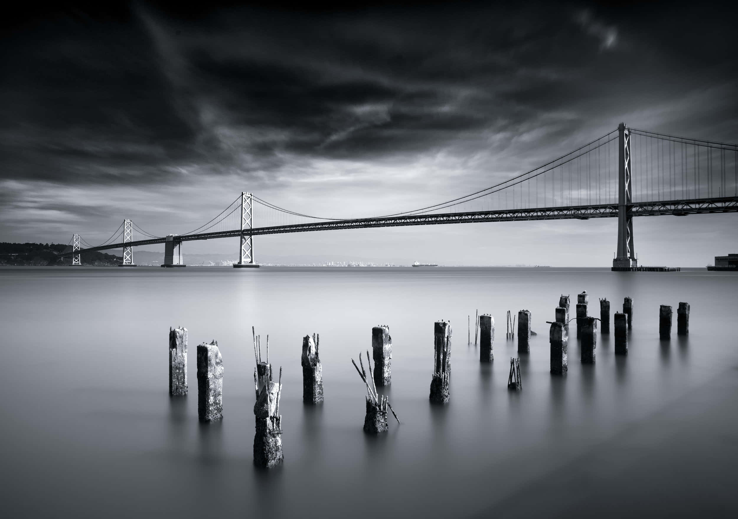 San Francisco Black And White 桌布 壁紙 — 海灣大橋的黑白長時間曝光照片，前景有風化的樁柱，背景是戲劇性的暴風雲。