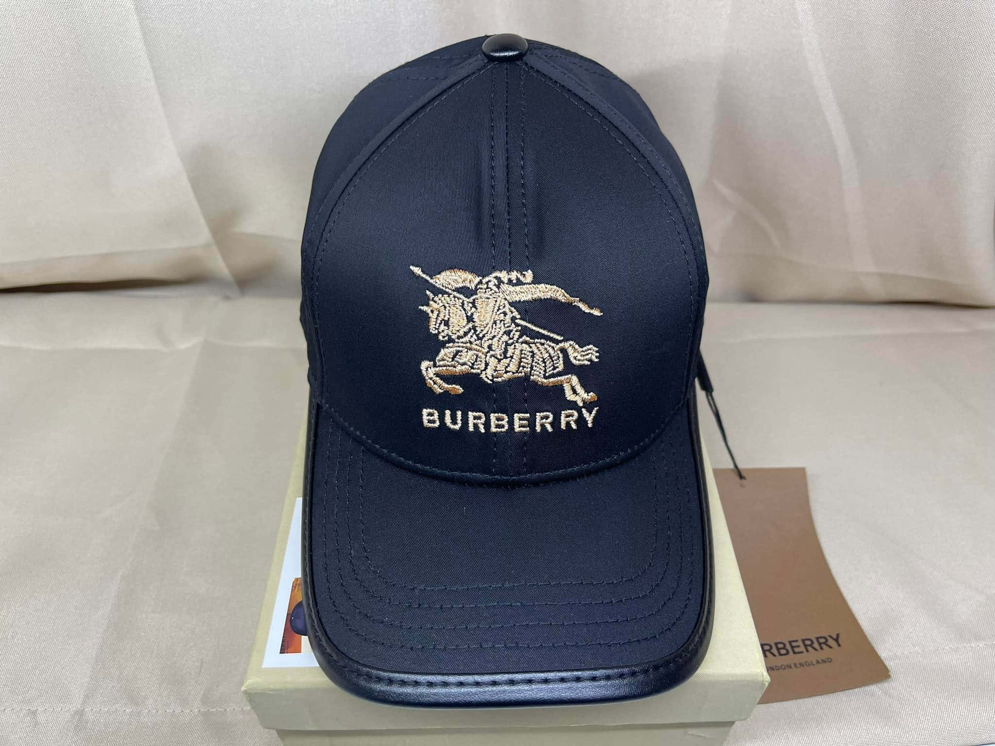 Burberry 桌布 壁紙 — 海軍藍色 Burberry 棒球帽，帶有金色刺繡騎士標誌，放在其品牌盒子上，背景為柔軟白色布料。