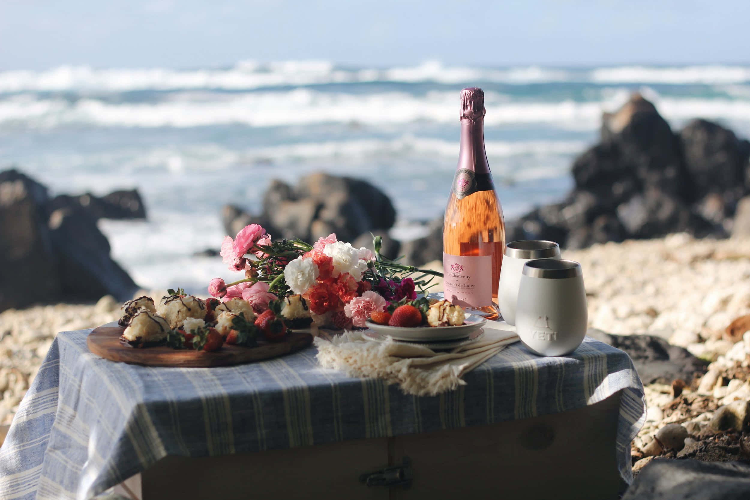 Beach Picnic 桌布 壁紙 — 海邊野餐佈置，在條紋布上擺放玫瑰酒、糕點和鮮花，旁邊是拍岸的浪花和岩石。