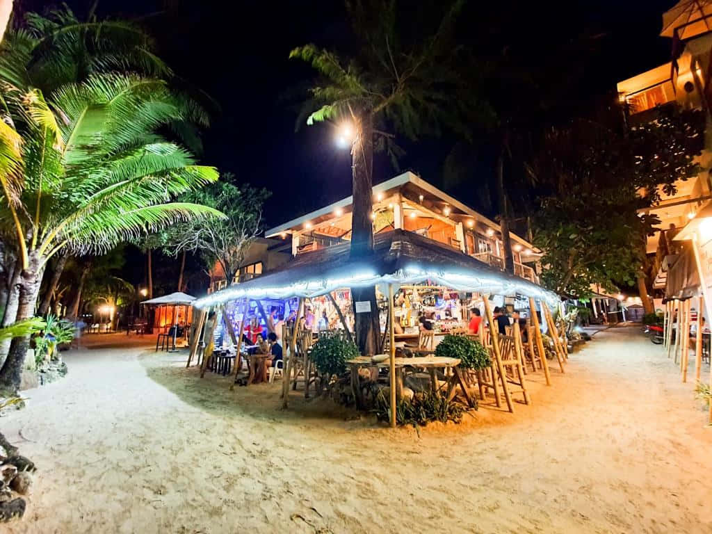 海灘 桌布 壁紙 — 海邊露天酒吧在夜晚棕櫚樹下溫暖地發光，客人在沙灘上享用飲品。