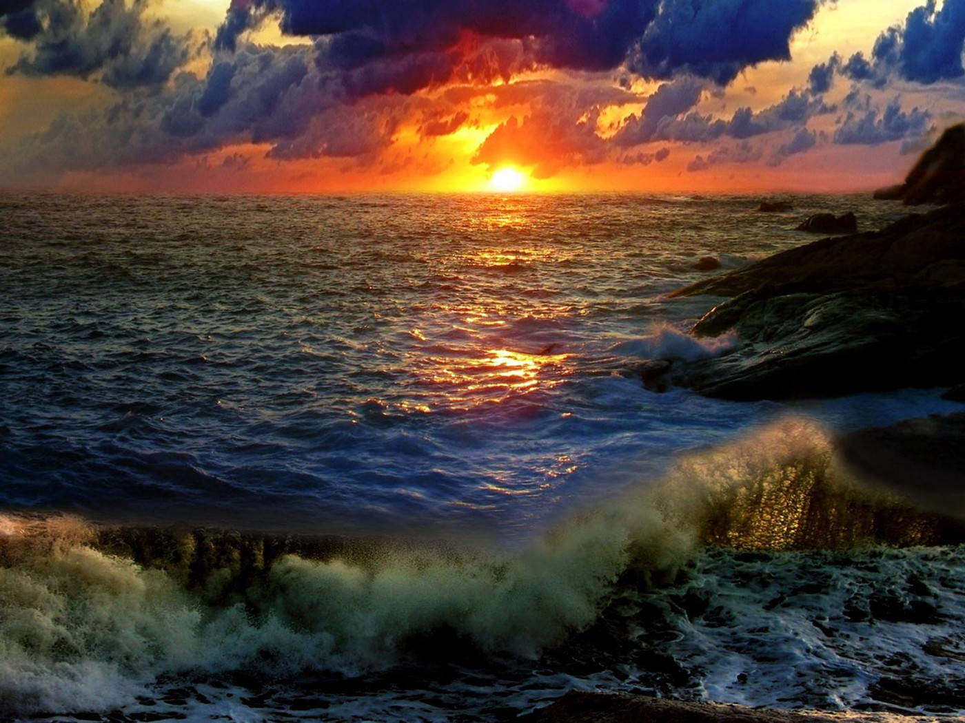 Sunset Aesthetic 桌布 壁紙 — 海面上戲劇性的日落，火紅的橙色太陽在洶湧的海浪上投下金色倒影，海浪拍打著黑暗的岩石海岸。
