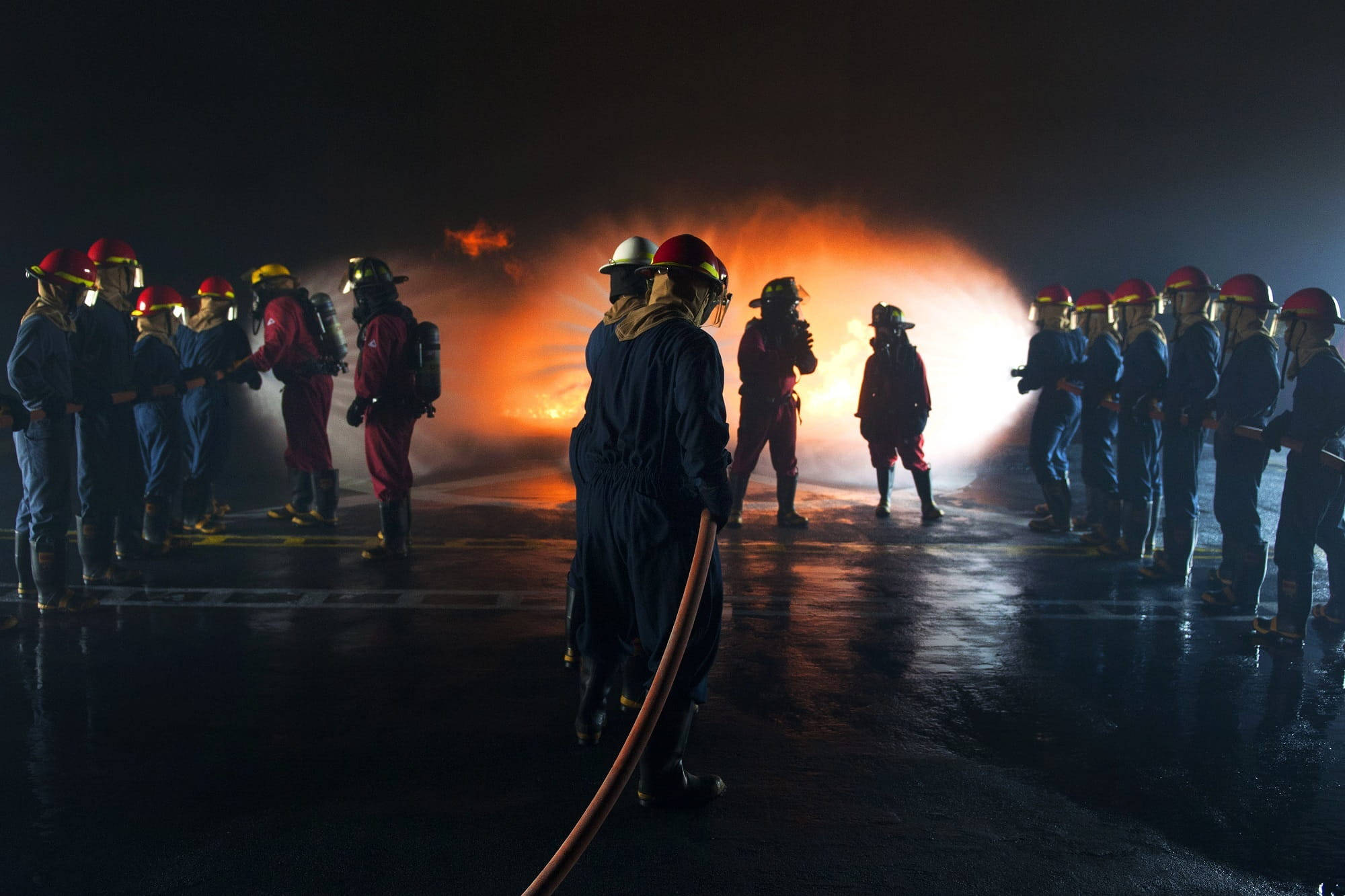 Firefighter 桌布 壁紙 — 消防員全副裝備在夜間列隊，身後發生大規模火災，在濕漉漉的路面上投射出橙色光芒。
