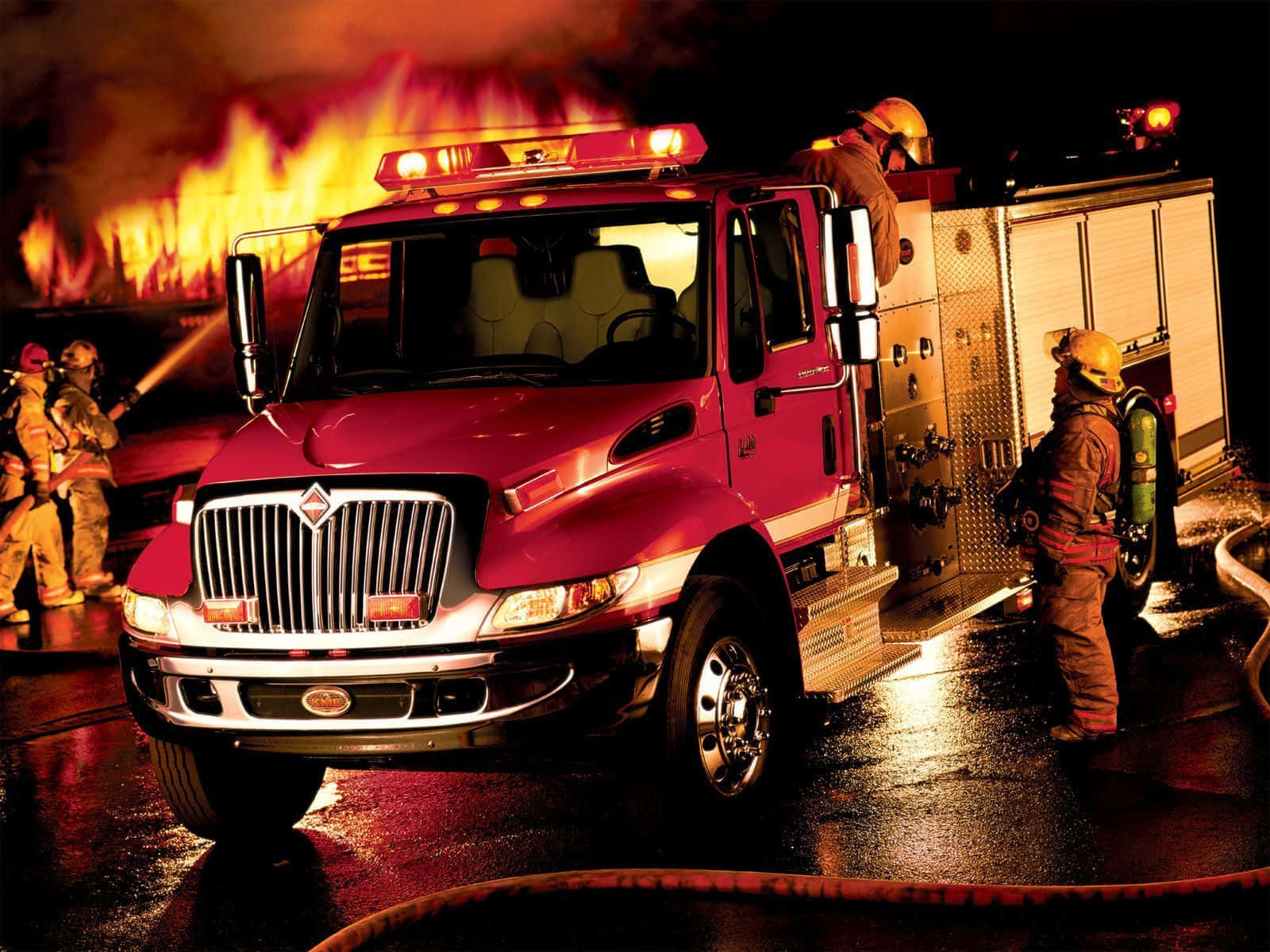 Fire Truck 桌布 壁紙 — 消防員在夜間用紅色消防車和軟管在熾熱的橙色光芒下撲滅猛烈的大火。