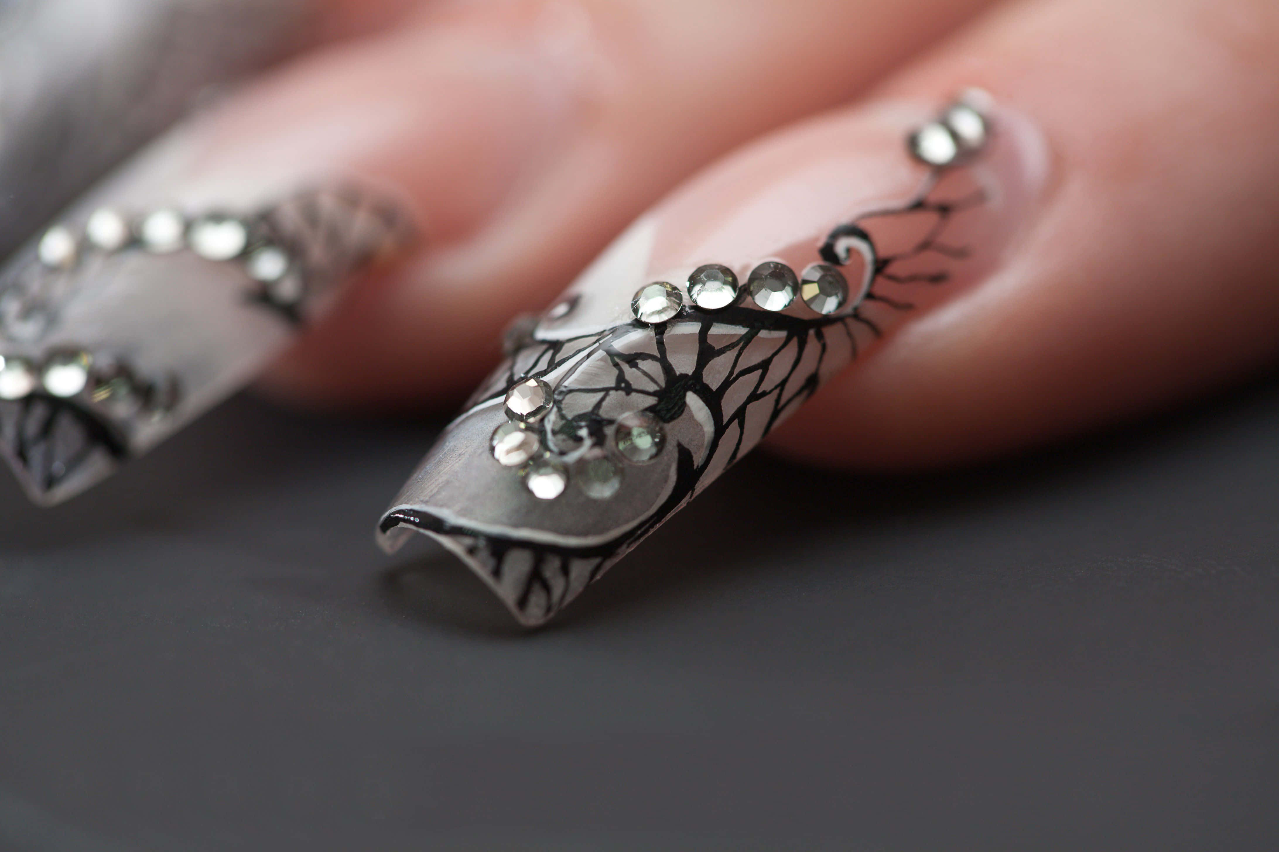 Cute Nails 桌布 壁紙 — 深灰色表面上帶有黑色樹枝圖案和閃亮水鑽的優雅裝飾指甲的特寫。
