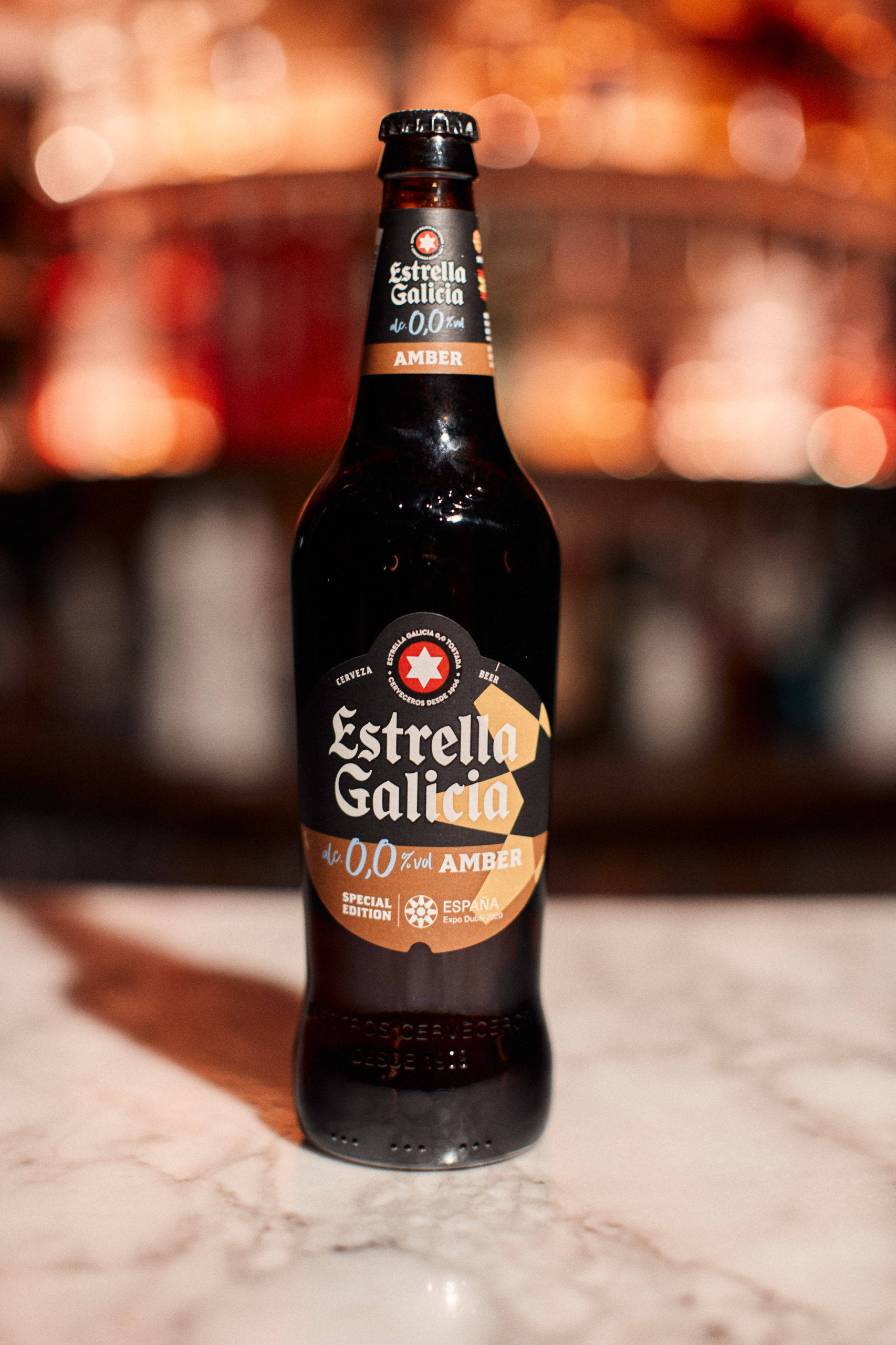 alcohol 桌布 壁紙 — 深琥珀色的Estrella Galicia 0.0啤酒瓶放在大理石吧檯上，背景有溫暖的散景燈光。