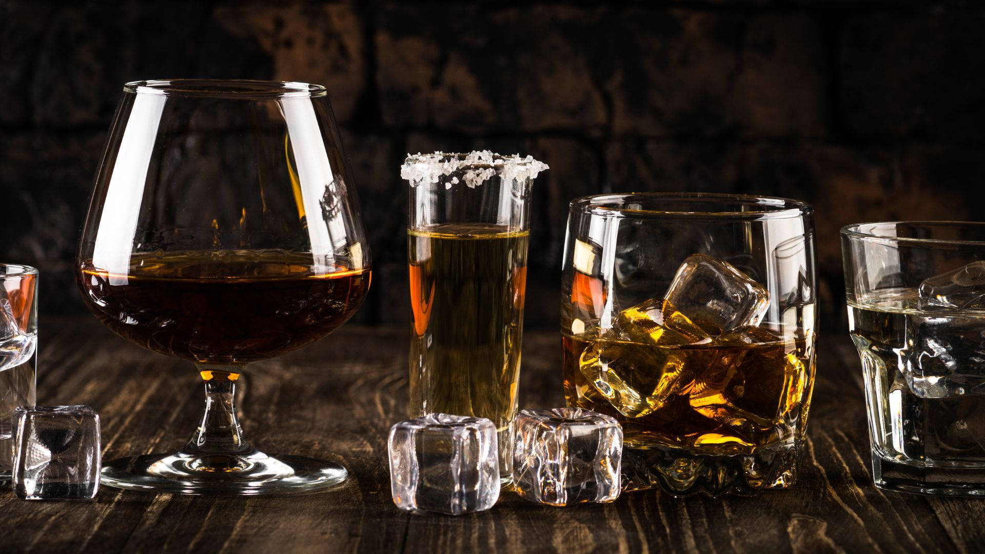 alcohol 桌布 壁紙 — 深琥珀色的烈酒和清澈的利口酒盛載於各種玻璃杯中，配上冰塊，放在質樸的木桌上，背景是帶紋理的磚牆。