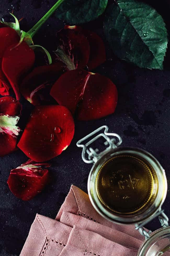 Rose Oil 桌布 壁紙 — 深紅色玫瑰花瓣和綠葉放在一個裝有金色液體的玻璃罐旁邊，置於帶有粉紅色布的深色紋理表面上。