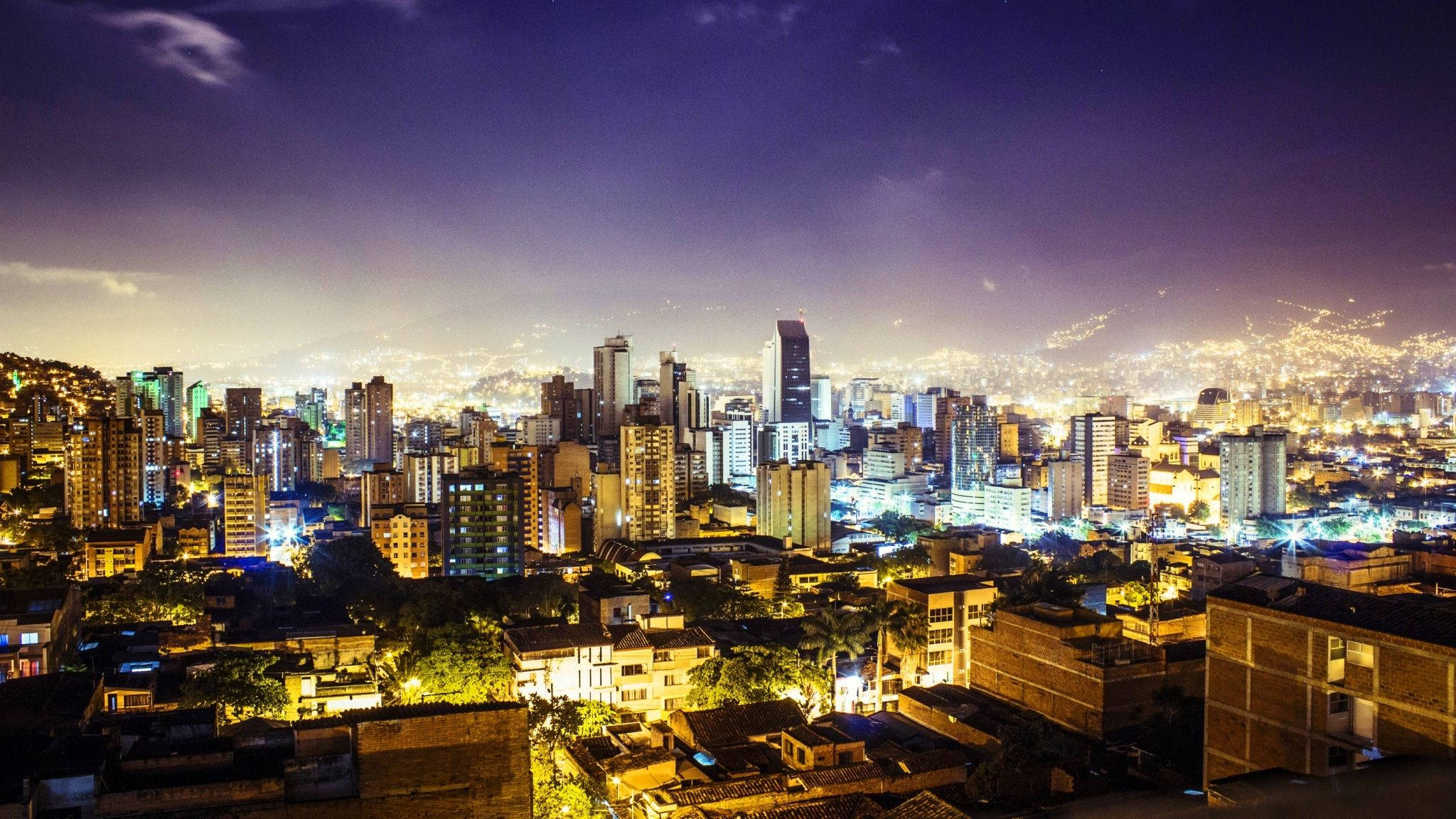 Colombia 桌布 壁紙 — 深紫色天空下，閃爍的摩天大樓和溫暖的路燈照亮了密集的城市社區，展現出廣闊的夜景。