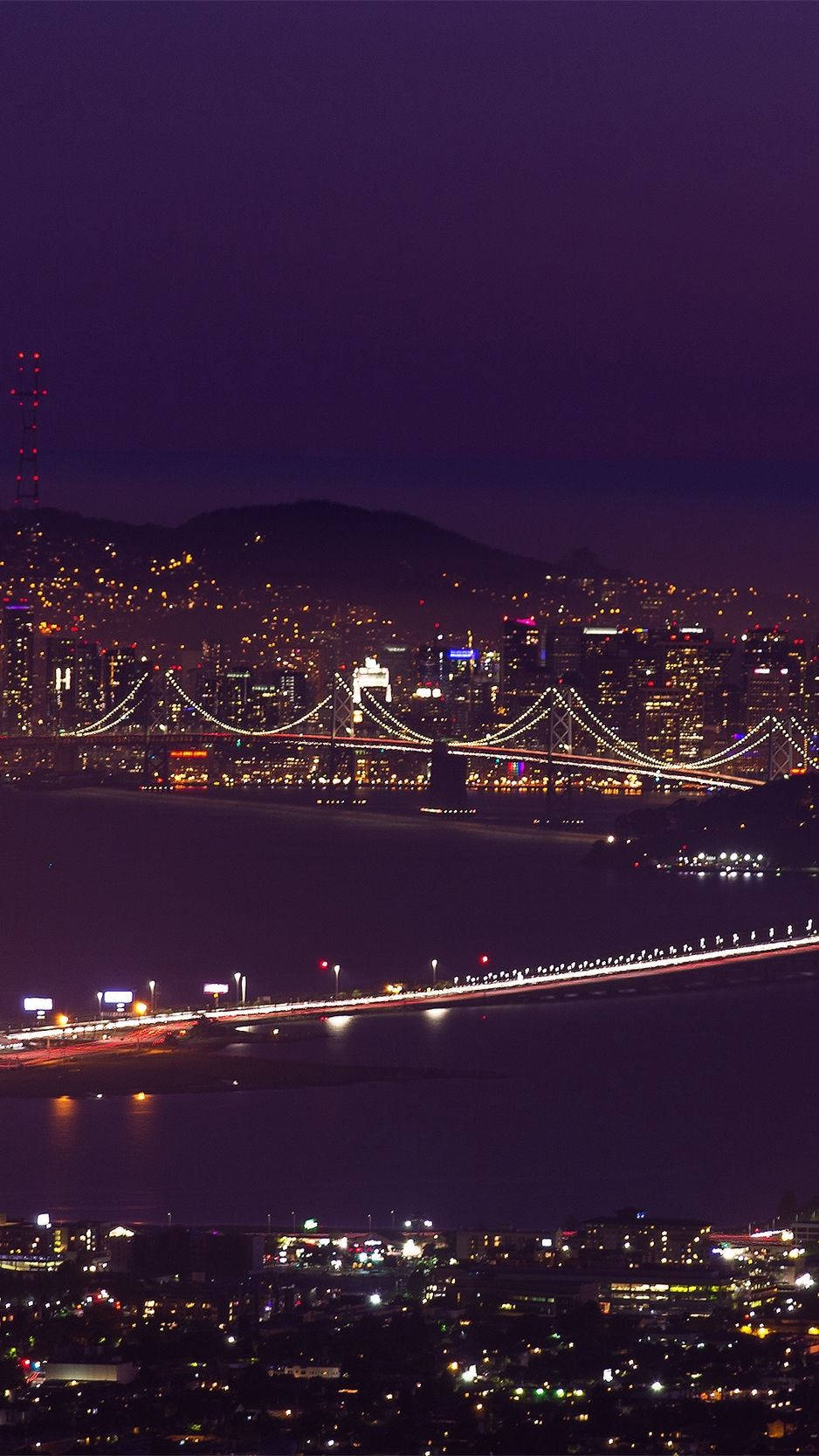 san francisco iPhone 桌布 壁紙 — 深紫色天空下，黑暗水面上閃爍著燈光的橋樑和遠山，構成發光的城市景觀的鳥瞰夜景。