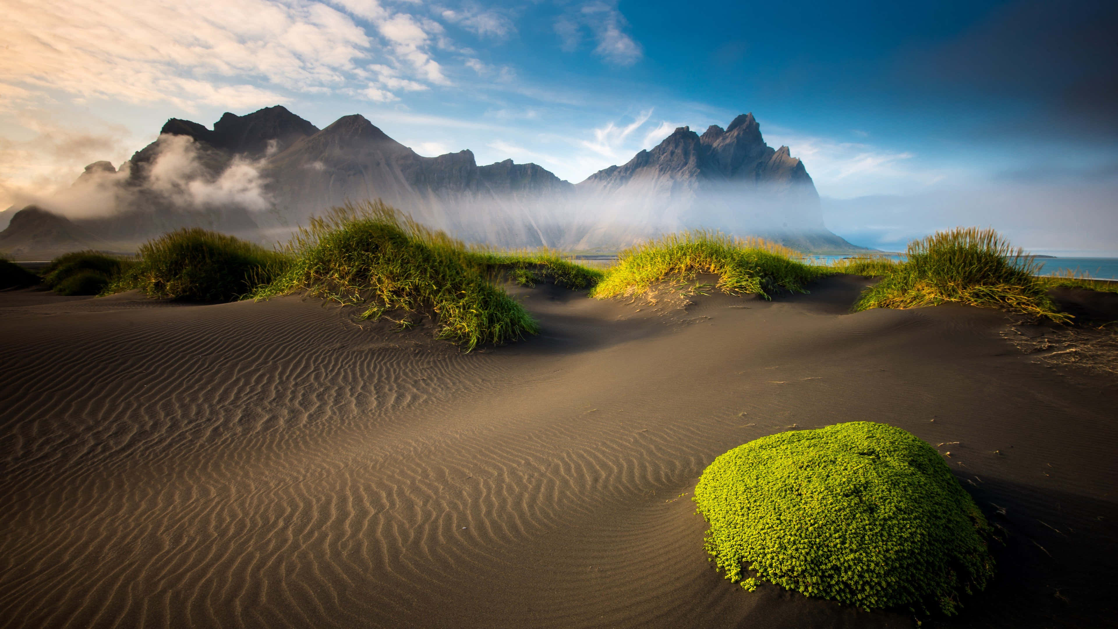 Iceland Desktop 桌布 壁紙 — 深色火山沙的漣漪在藍天白雲和金色雲彩下延伸至霧氣繚繞的山巒。
