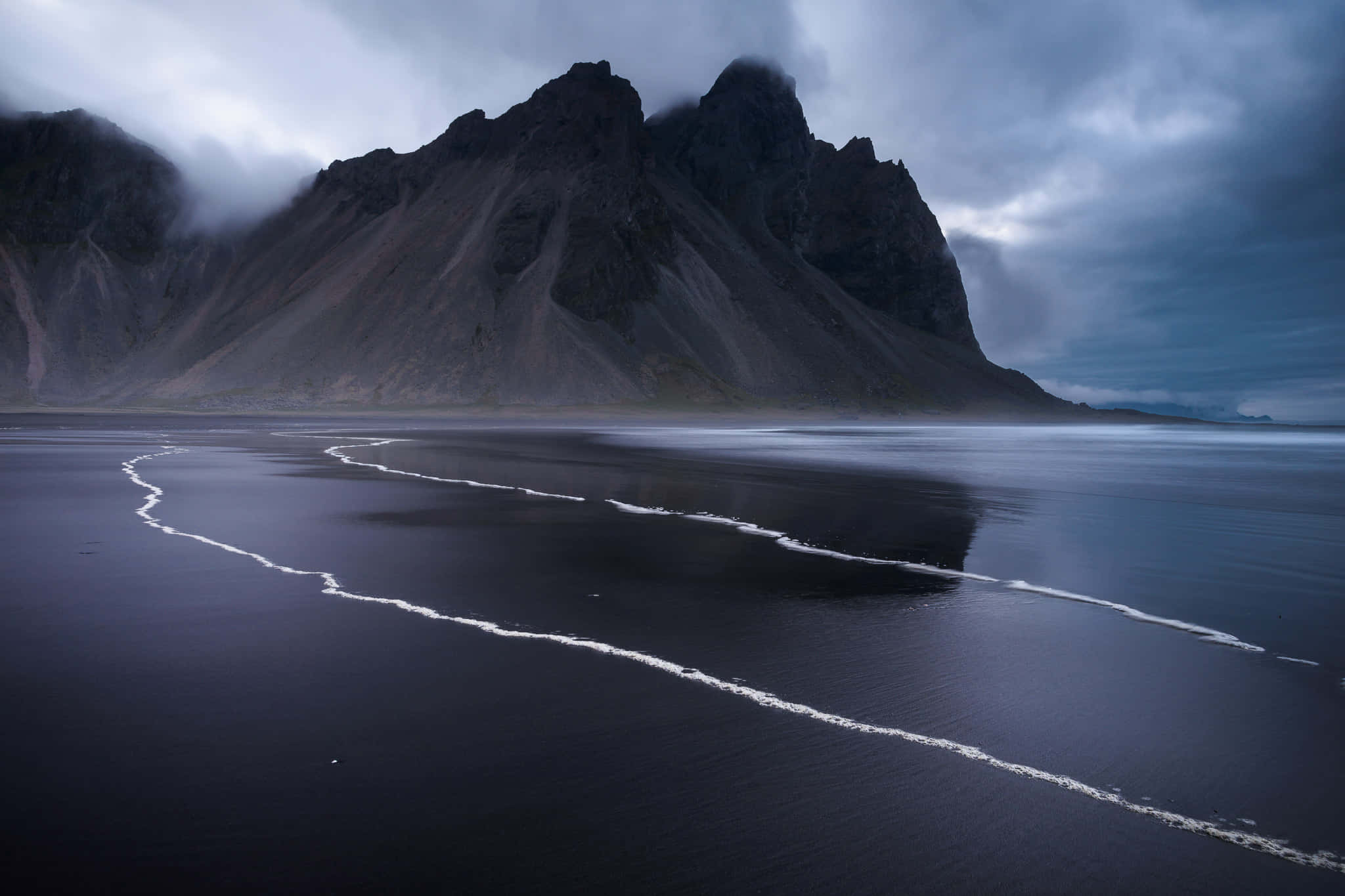 Google Landscape 桌布 壁紙 — 深色火山海灘，溫柔的海浪在陰沉多雲的天空下留下白色泡沫痕跡，背景中鋸齒狀的山脈若隱若現。