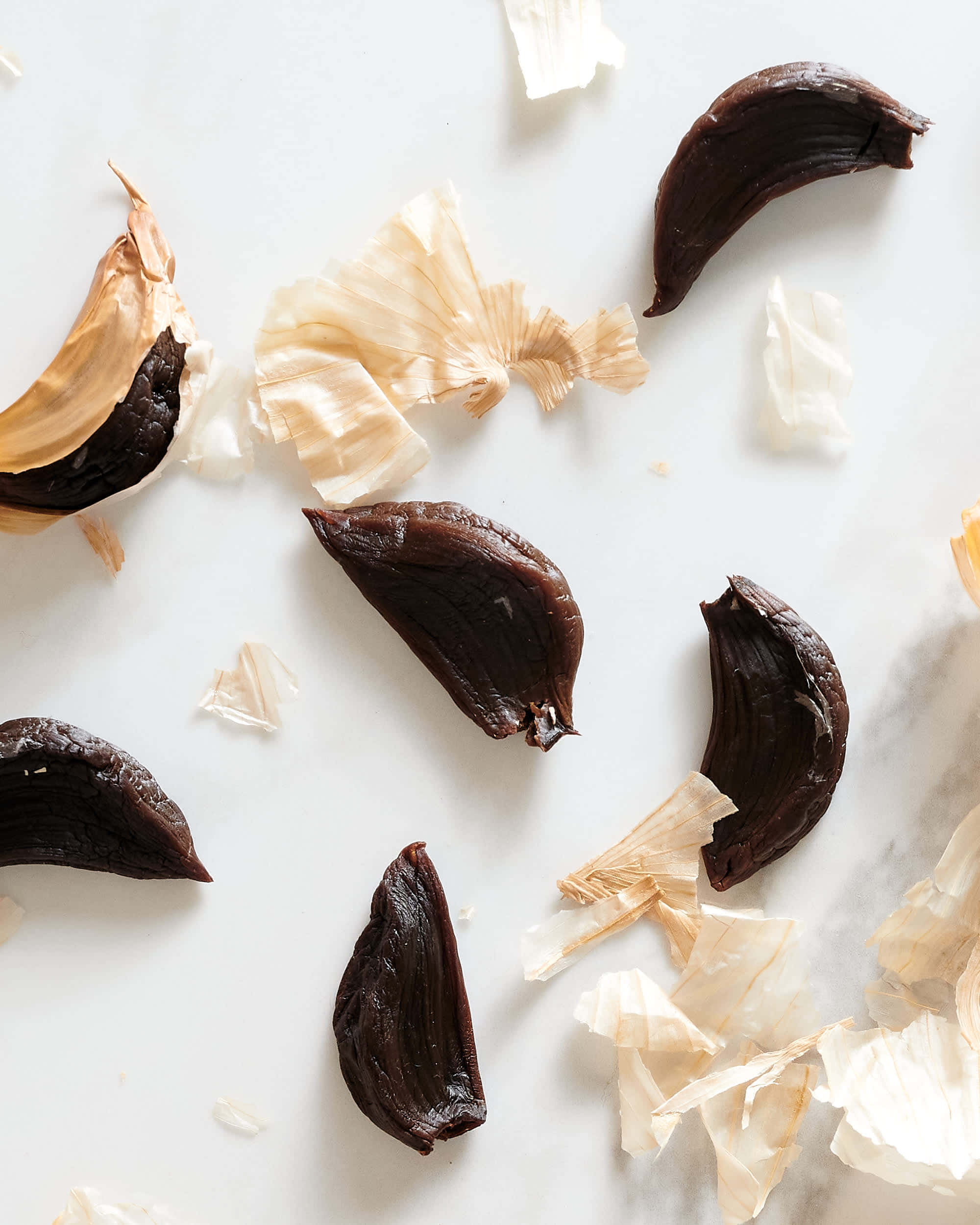 Black Garlic 桌布 壁紙 — 深色烤蒜瓣和紙狀蒜皮散落在白色大理石表面上。