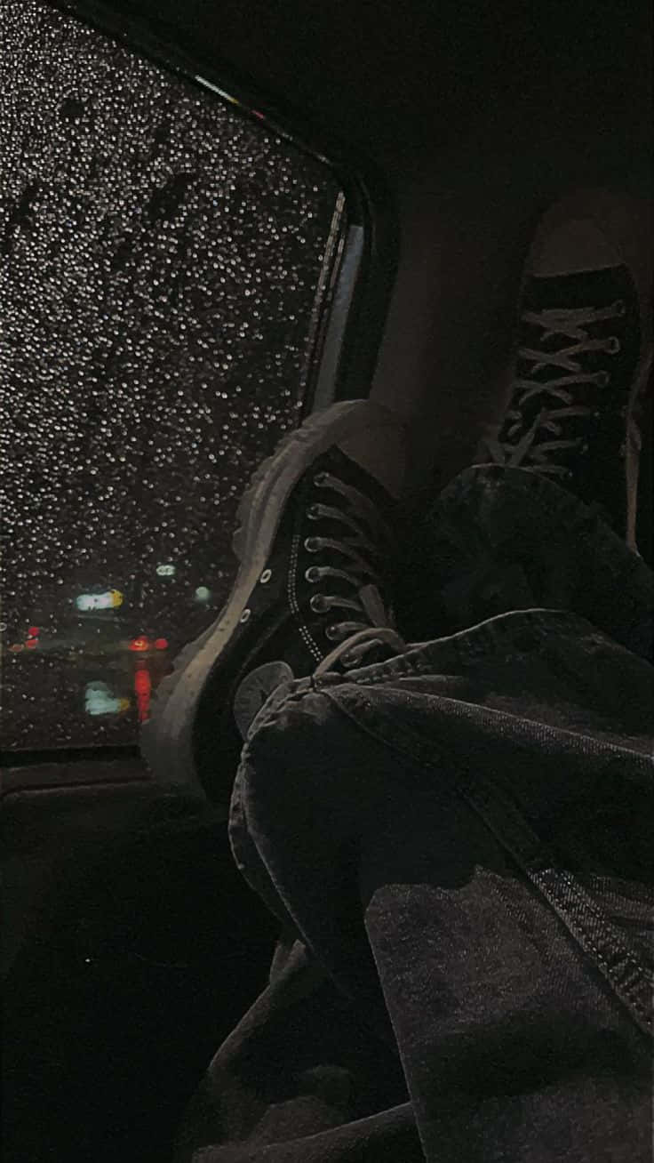 垃圾美學 桌布 壁紙 — 深色牛仔褲腿和黑色運動鞋在車內休息，雨滴劃過車窗，顯露出外面模糊的城市燈光。