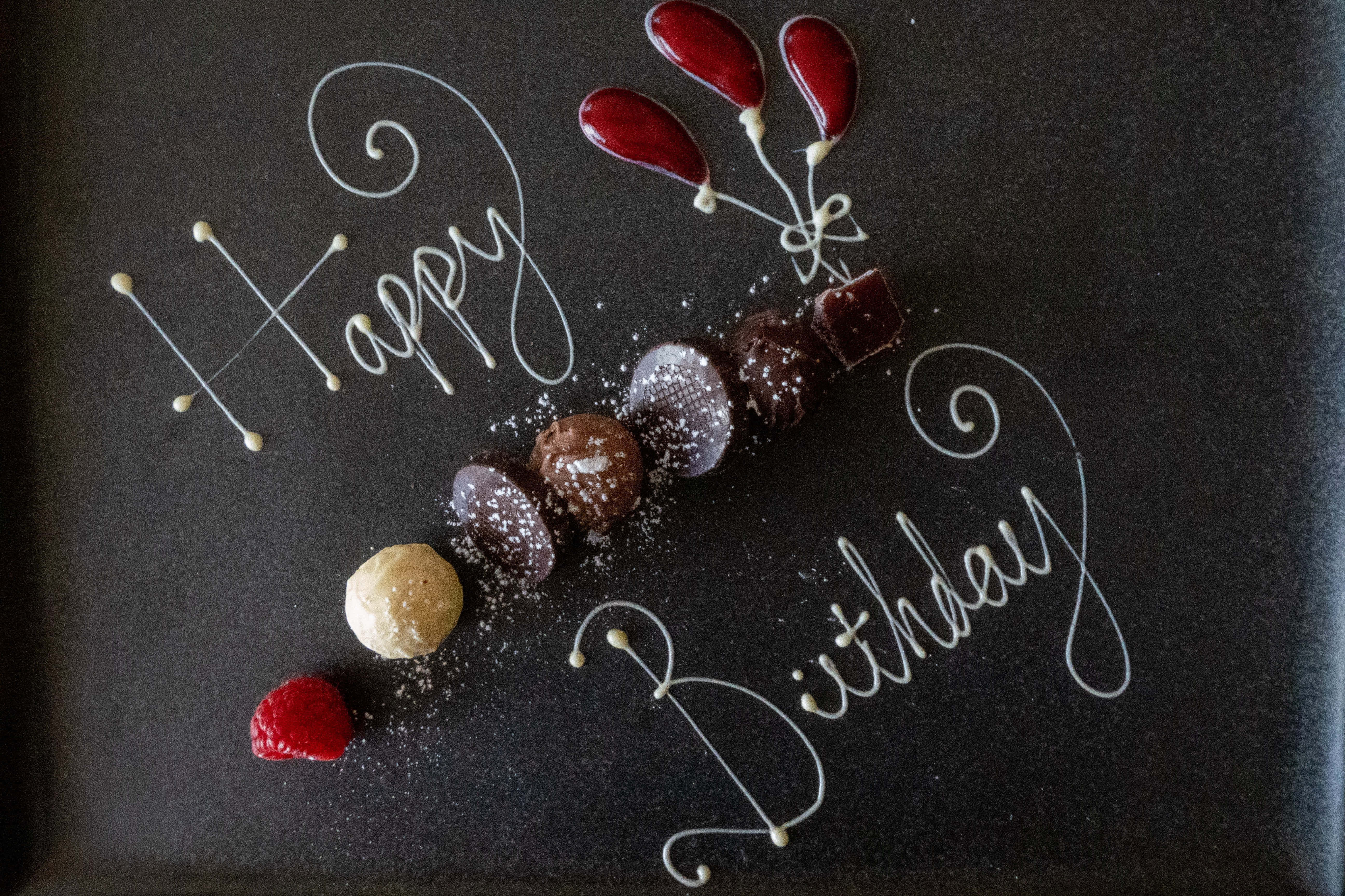 Cute Birthday 桌布 壁紙 — 深色盤子上以白巧克力寫著「Happy Birthday」，搭配紅色氣球裝飾和各種撒上糖粉的松露巧克力。