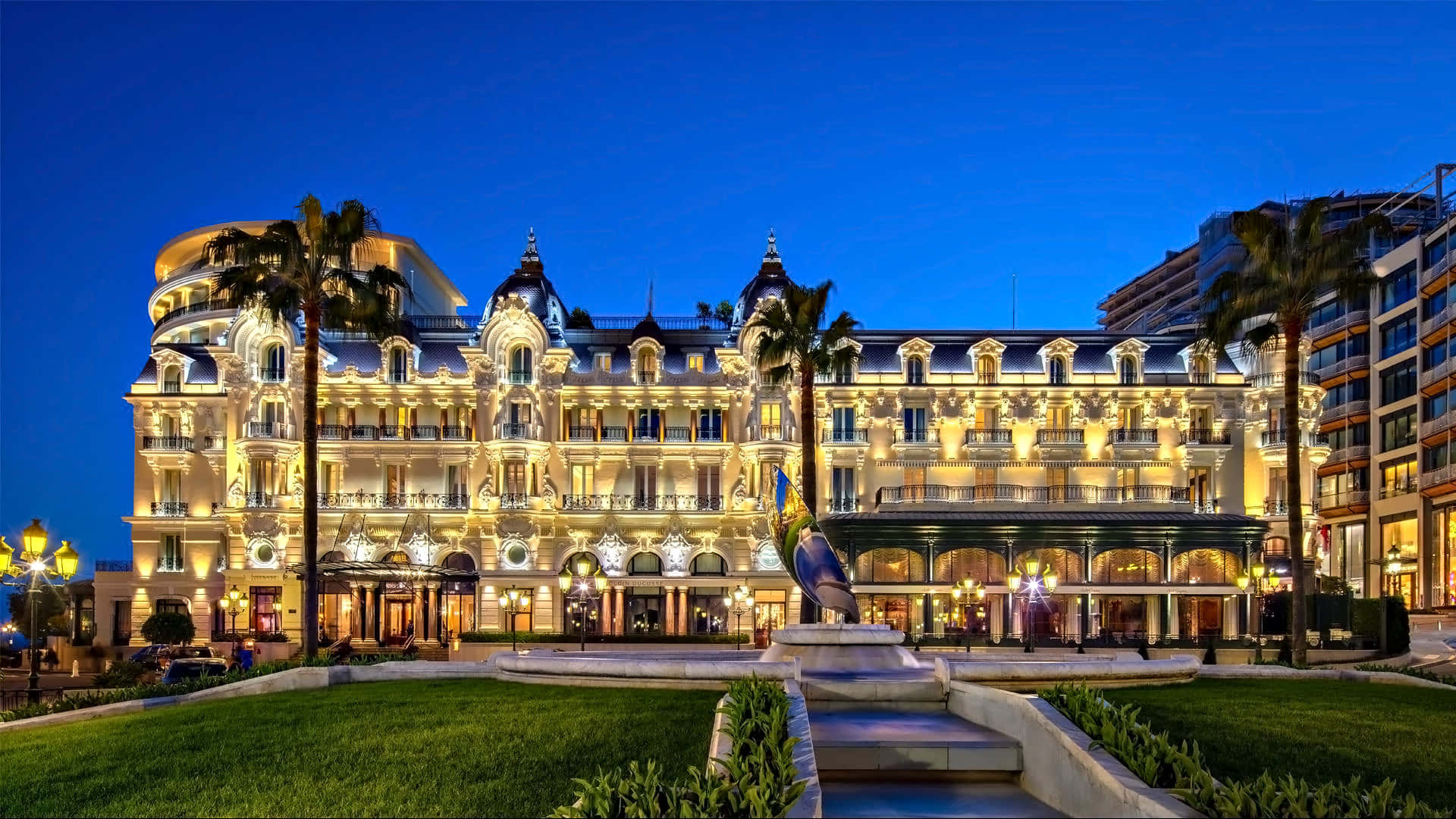Monaco 桌布 壁紙 — 深藍天空下，暮色中宏偉的燈光酒店外牆，棕櫚樹和修剪整齊的草坪。
