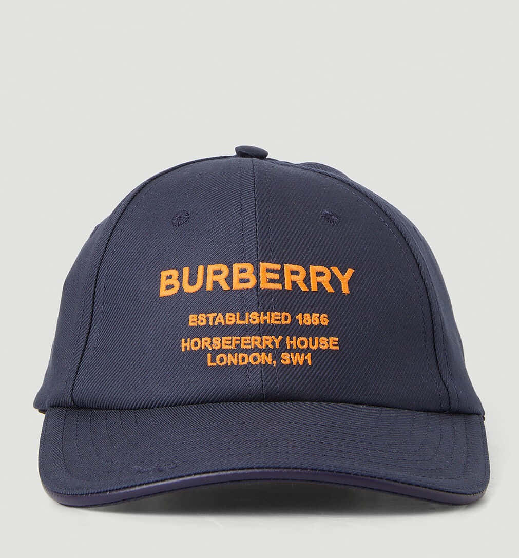 Burberry 桌布 壁紙 — 深藍色 Burberry 棒球帽，帶有橙色刺繡文字，詳細說明其傳統和倫敦地址，背景為純色。