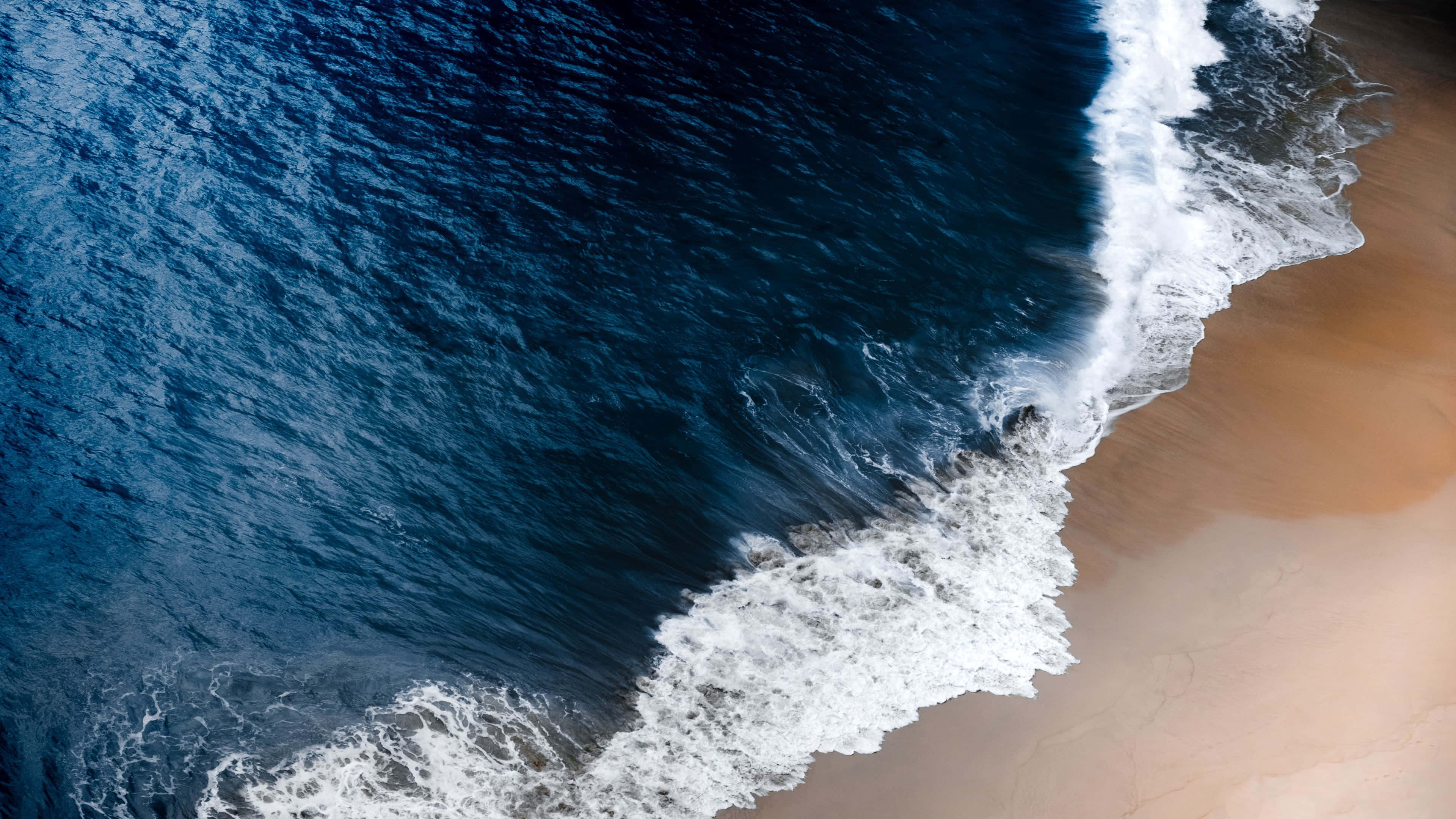 Beach Waves 桌布 壁紙 — 深藍色海浪拍打金色沙灘並泛起白色泡沫的航拍視角。