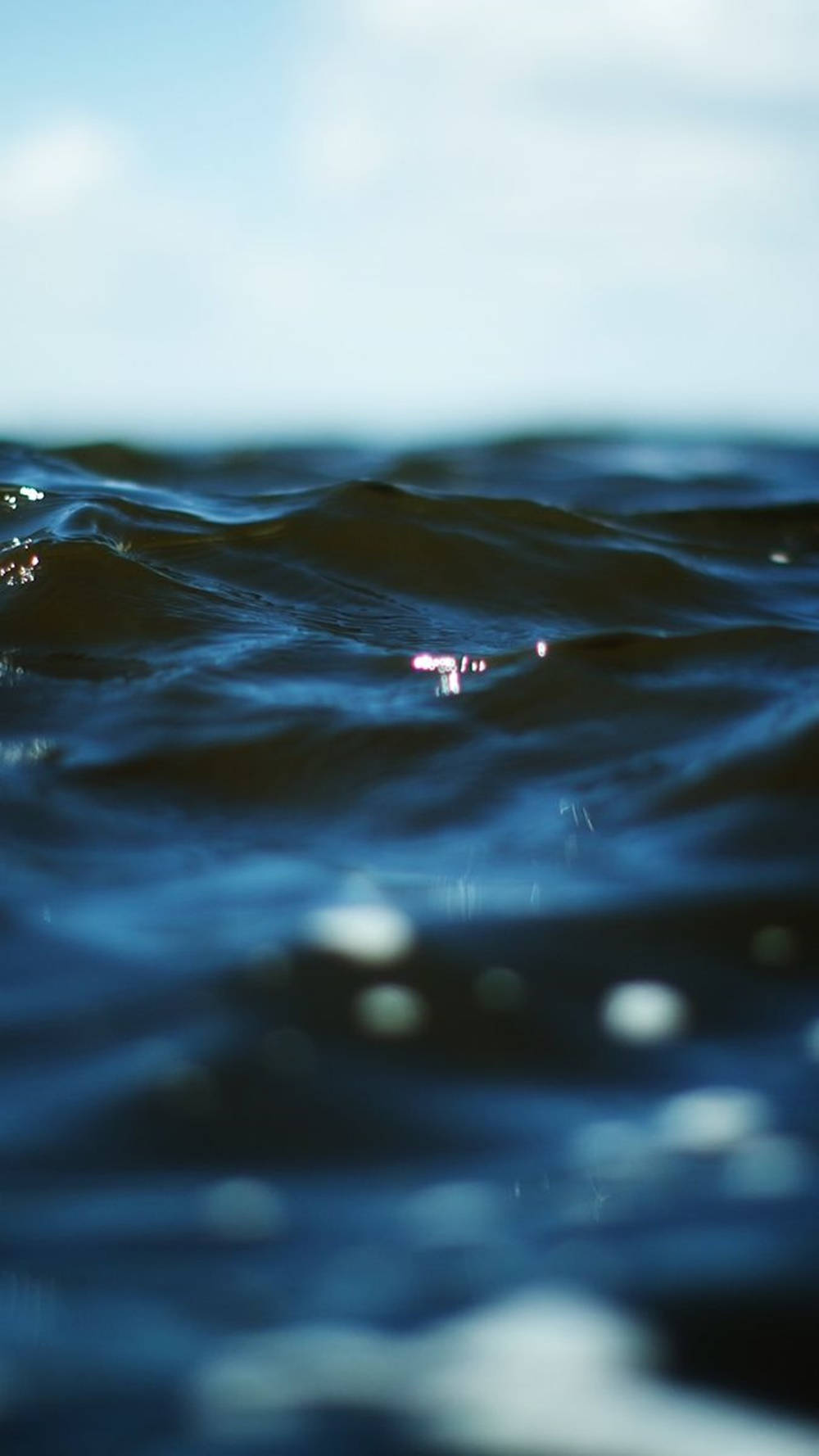 Ocean iPhone 桌布 壁紙 — 深藍色海浪的特寫，帶有陽光閃爍和柔和模糊的天空背景。