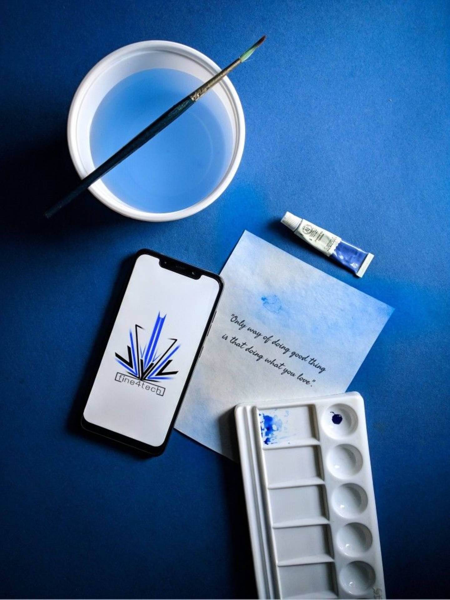 藍色美學 桌布 壁紙 — 深藍色表面上的藝術用品平鋪擺設，包括畫筆、水杯、調色板和電話。