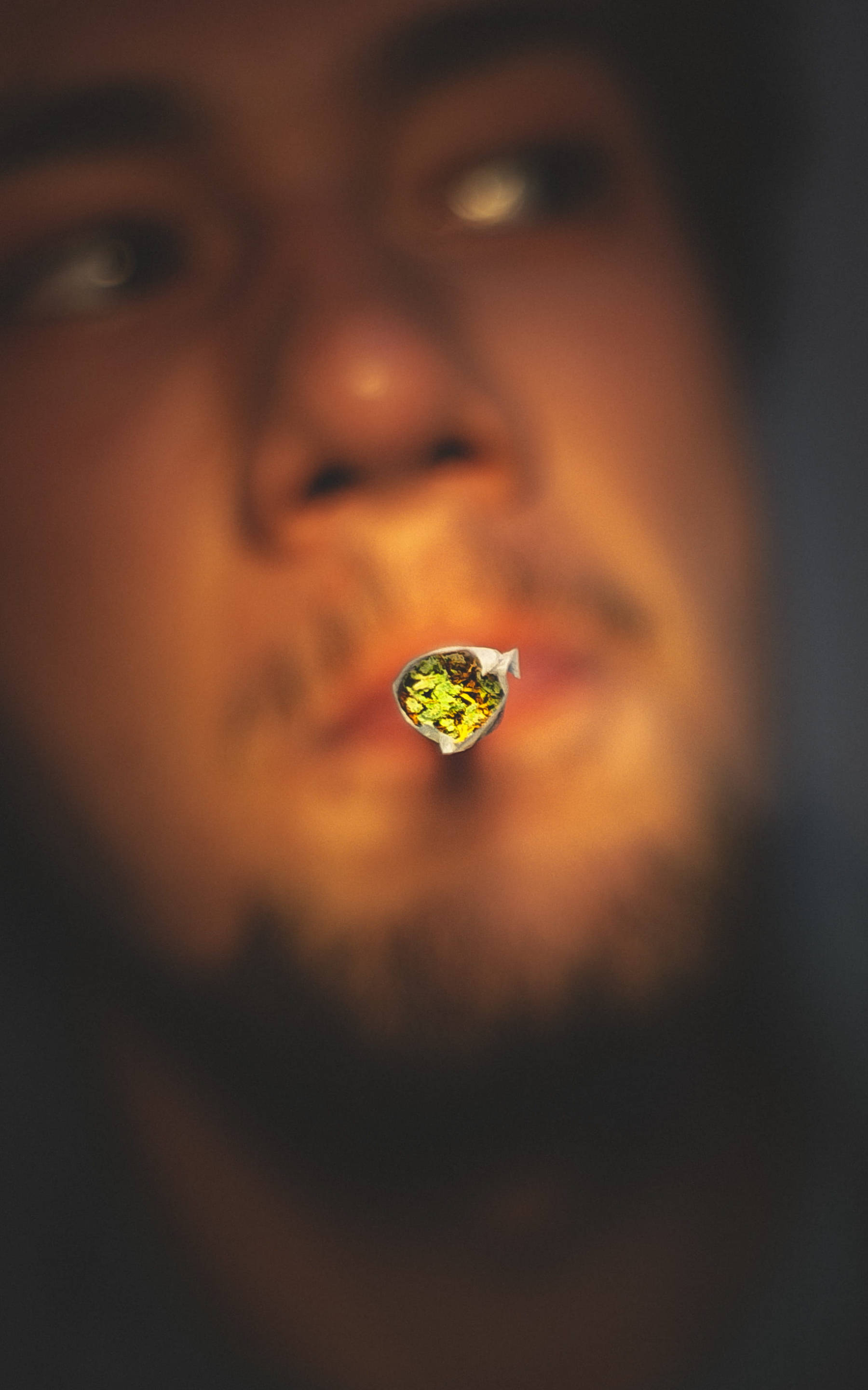 吸大麻 桌布 壁紙 — 淺景深將焦點銳利地對準在嘴唇之間的一根點燃的大麻煙上，而背景中模糊的男性面孔溫暖地發光。