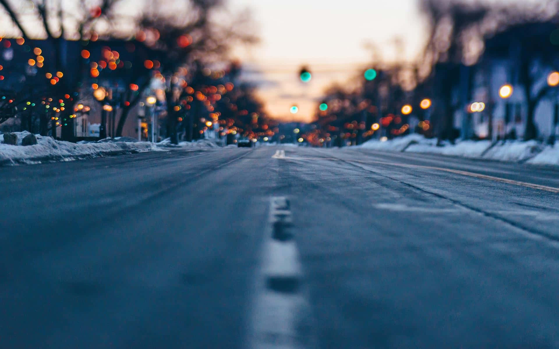 Road 桌布 壁紙 — 淺景深捕捉了黃昏時分積雪的都市街道，模糊的暖色燈光和光禿禿的樹木排列在路邊。
