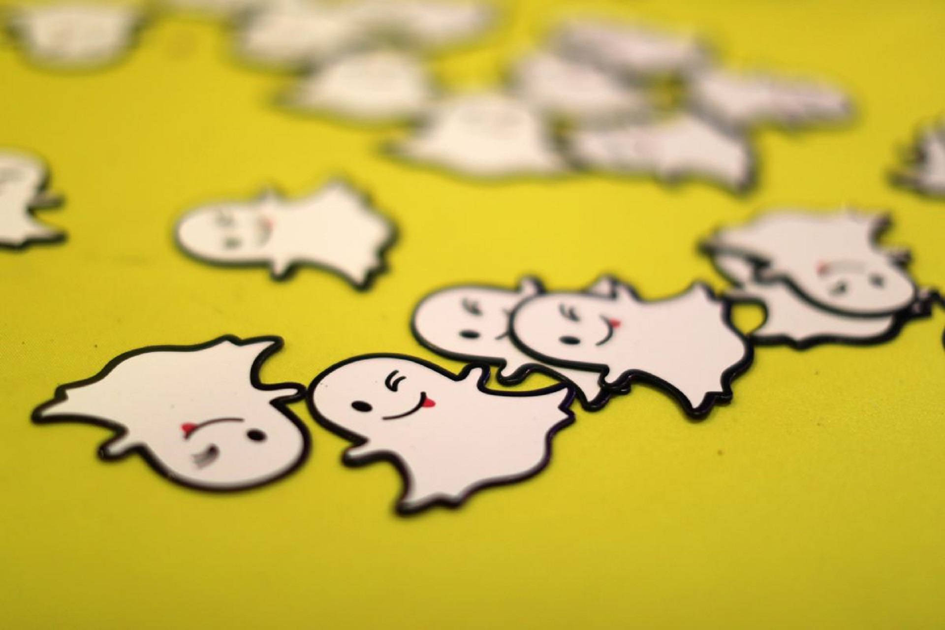 Snapchat 桌布 壁紙 — 淺景深突出了一組微笑的白色鬼魂形狀貼紙，帶有黑色輪廓，位於充滿活力的黃色表面上。