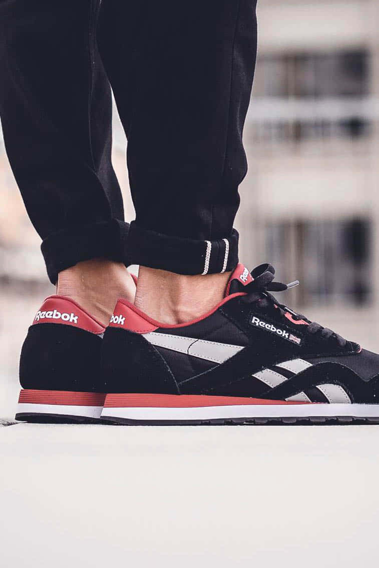 銳步 桌布 壁紙 — 淺灰色表面上，黑色、紅色和白色 Reebok 運動鞋搭配捲邊黑色褲子的特寫。