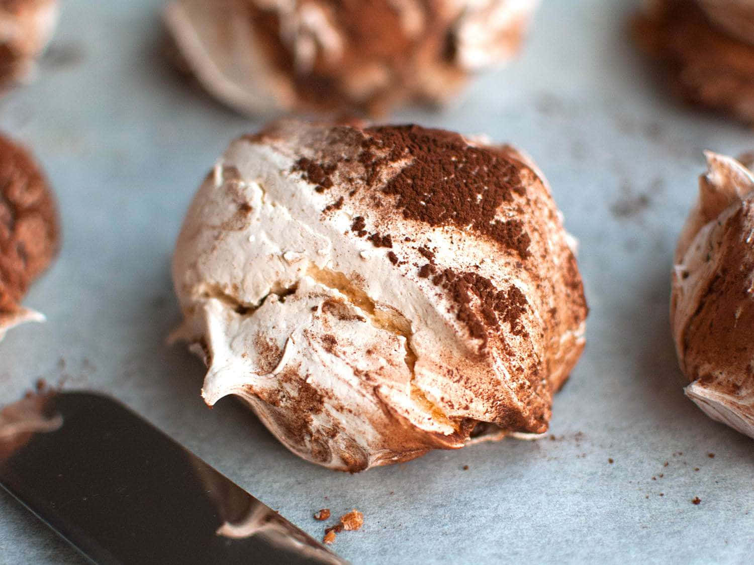 Meringue 桌布 壁紙 — 淺灰色表面上撒有可可粉的金黃色蛋白脆餅特寫，附近有一把刮刀。