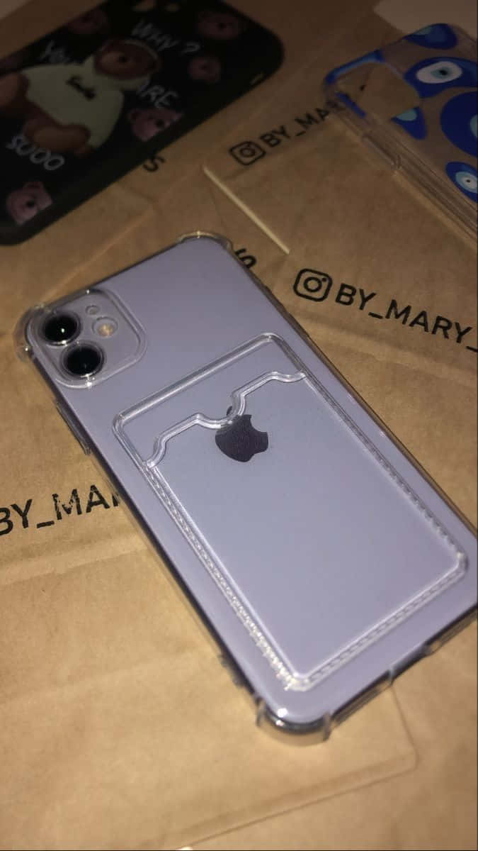 iPhone 12 桌布 壁紙 — 淺紫色iPhone放在透明手機殼（附卡槽）中，擱在印有BY_MARY品牌的棕色紙上。