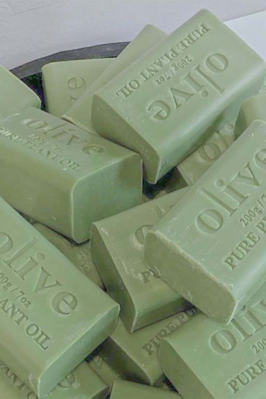 Aesthetic Sage Green 桌布 壁紙 — 淺綠色橄欖油肥皂條堆疊在一起，帶有浮雕文字，放在一個深色碗中，背景為白色。