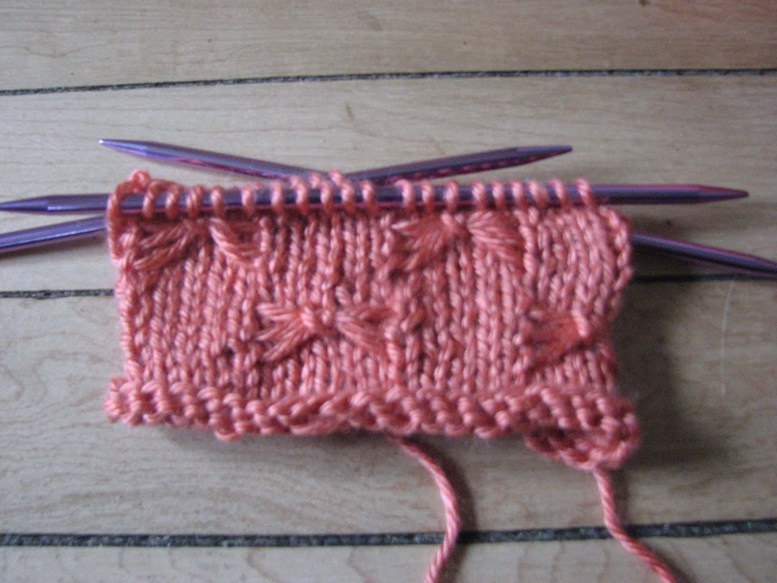 Butterfly Stitch 桌布 壁紙 — 淺色木面上紫色編織針上帶有葉子圖案的珊瑚粉紅色針織布料的特寫。