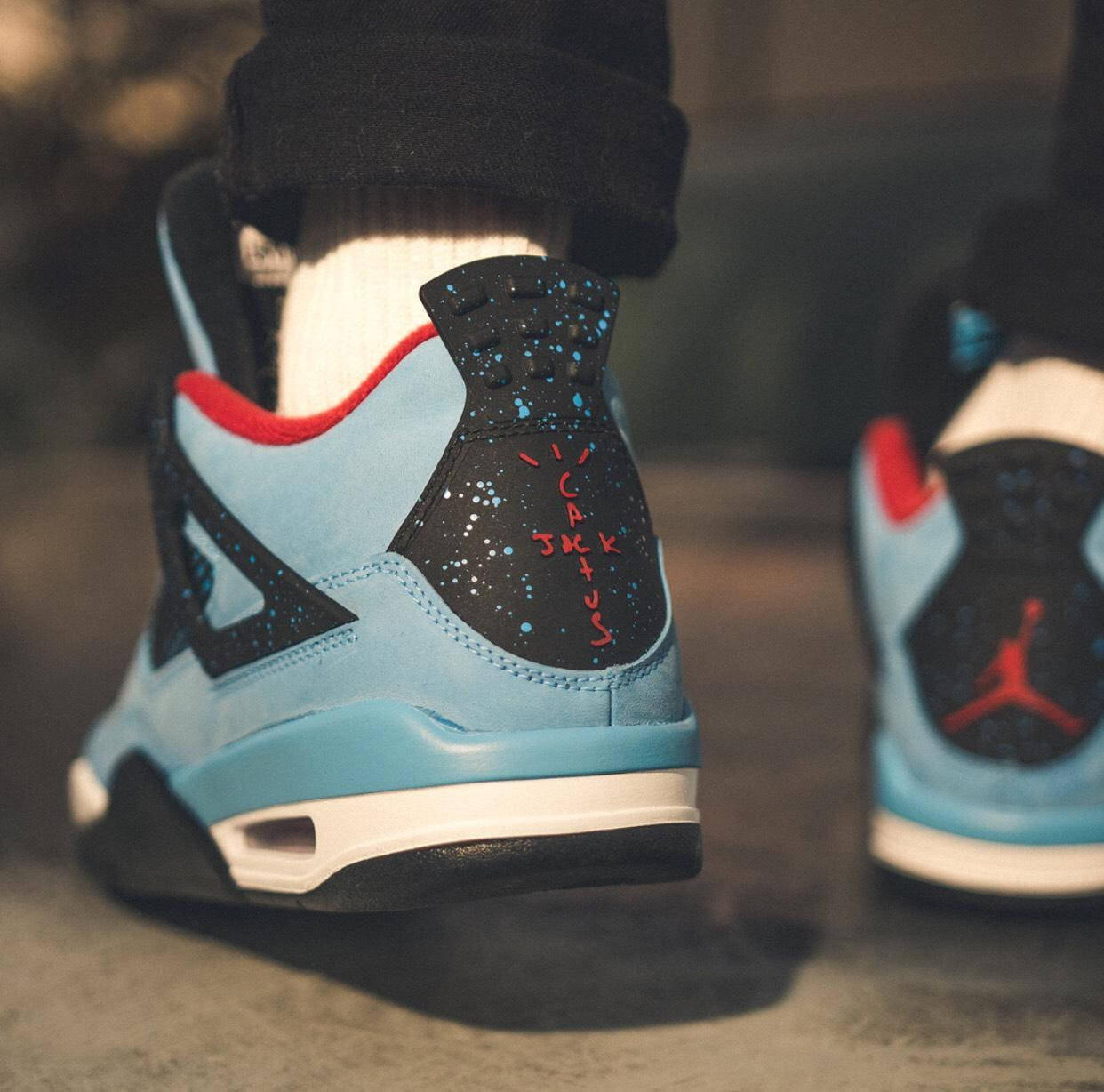 崔維斯·史考特 桌布 壁紙 — 淺藍、黑色和紅色 Air Jordan 4 運動鞋的特寫，鞋跟帶有 'YIKES' 塗鴉，背景為模糊的城市地板。