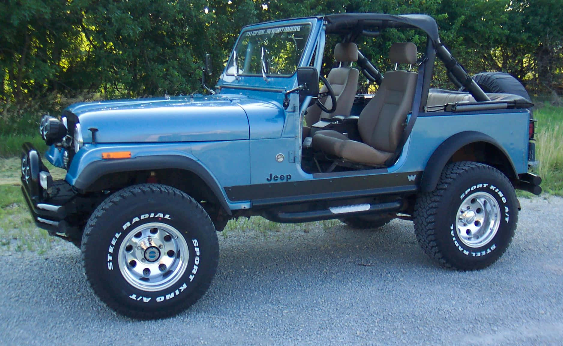 Jeep Cj 桌布 壁紙 — 淺藍色Jeep Wrangler，配備米色內裝及大型越野輪胎，停放在碎石路上，附近有綠樹。
