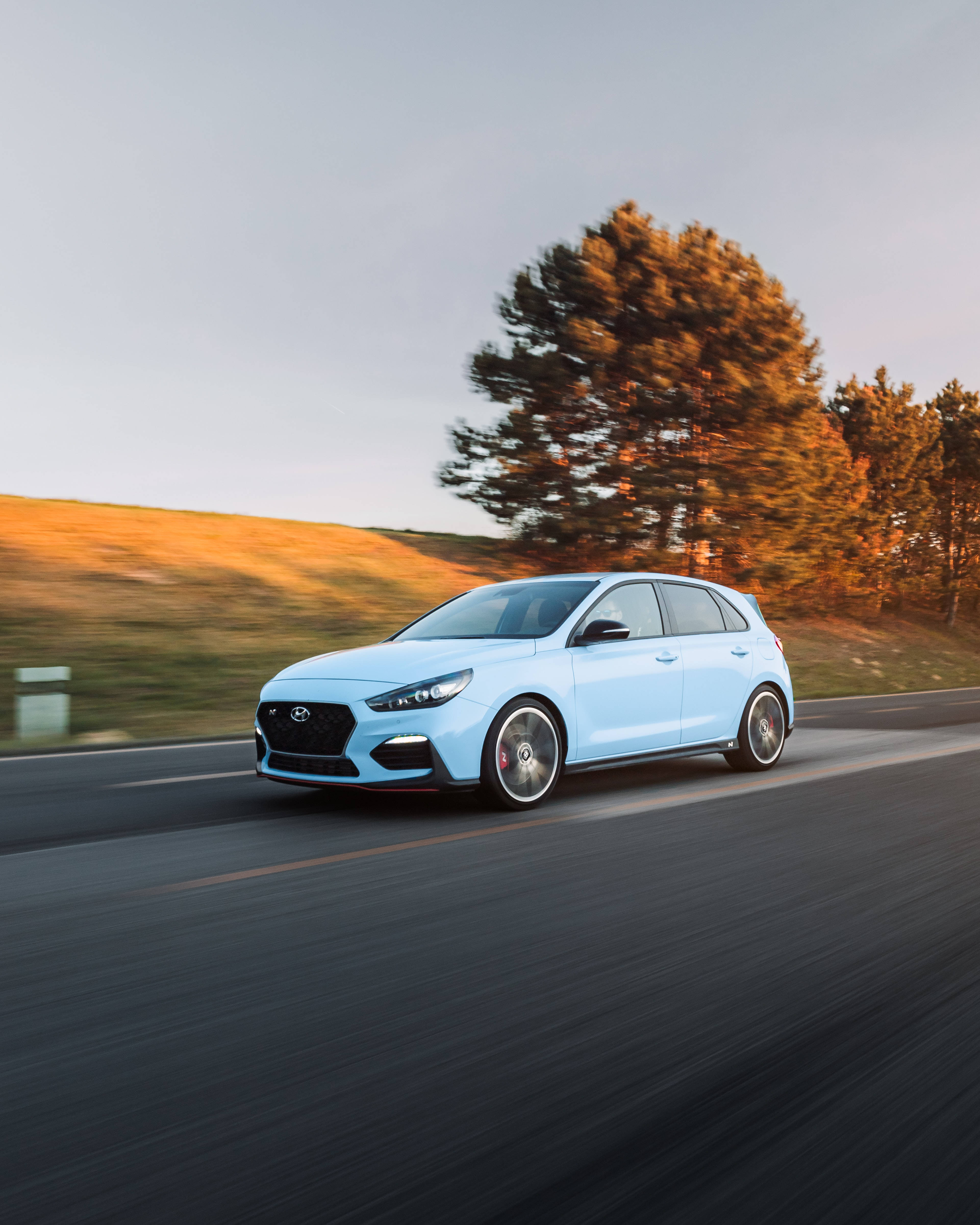 Hyundai 桌布 壁紙 — 淺藍色現代 i30 N 掀背車在日落時分沿著蜿蜒的道路疾馳，金色樹木和模糊動感。