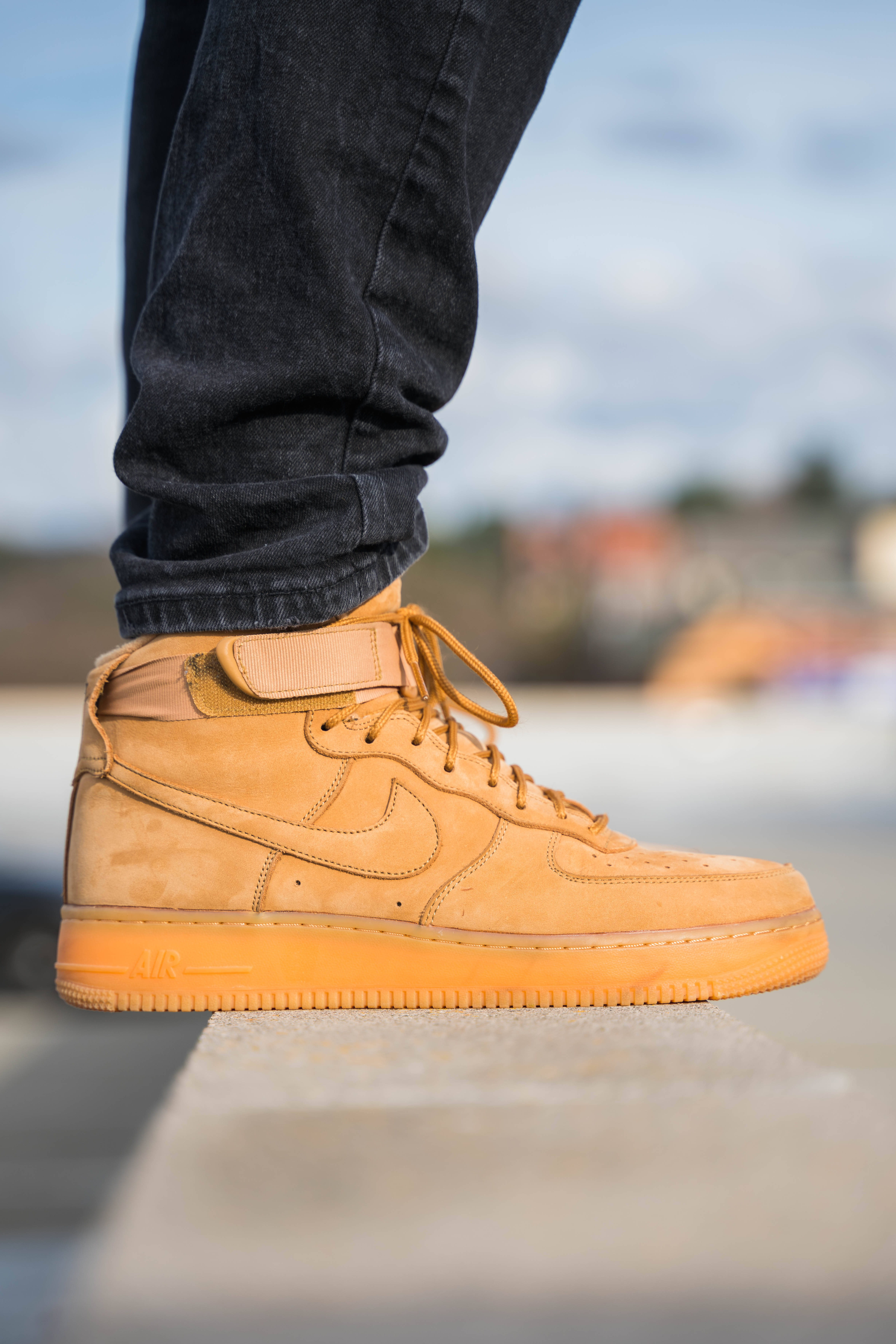 Sneaker 桌布 壁紙 — 淺褐色麂皮Nike Air Force 1運動鞋搭配深色牛仔褲，站在混凝土邊緣上，背景是模糊的城市景觀的近距離特寫。