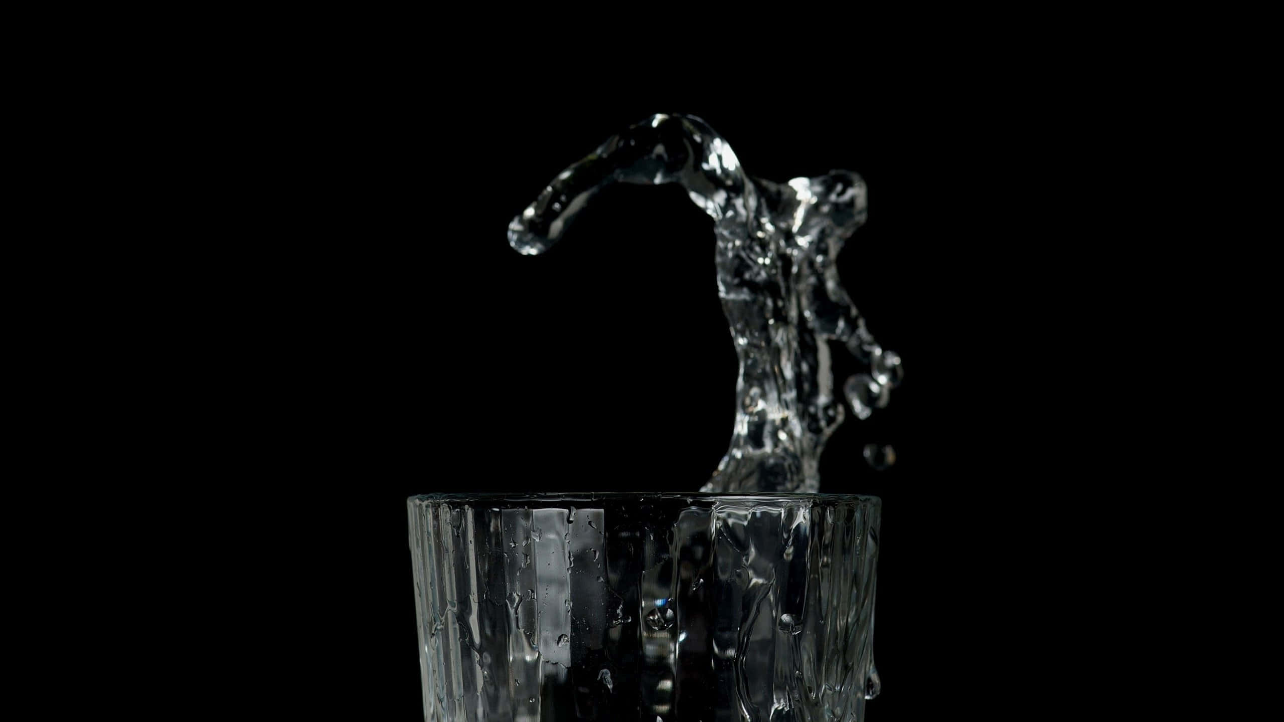 Glass Of Water 桌布 壁紙 — 清澈的水從有紋理的玻璃杯中向上拱起，形成動態的水花，背景是純黑色。