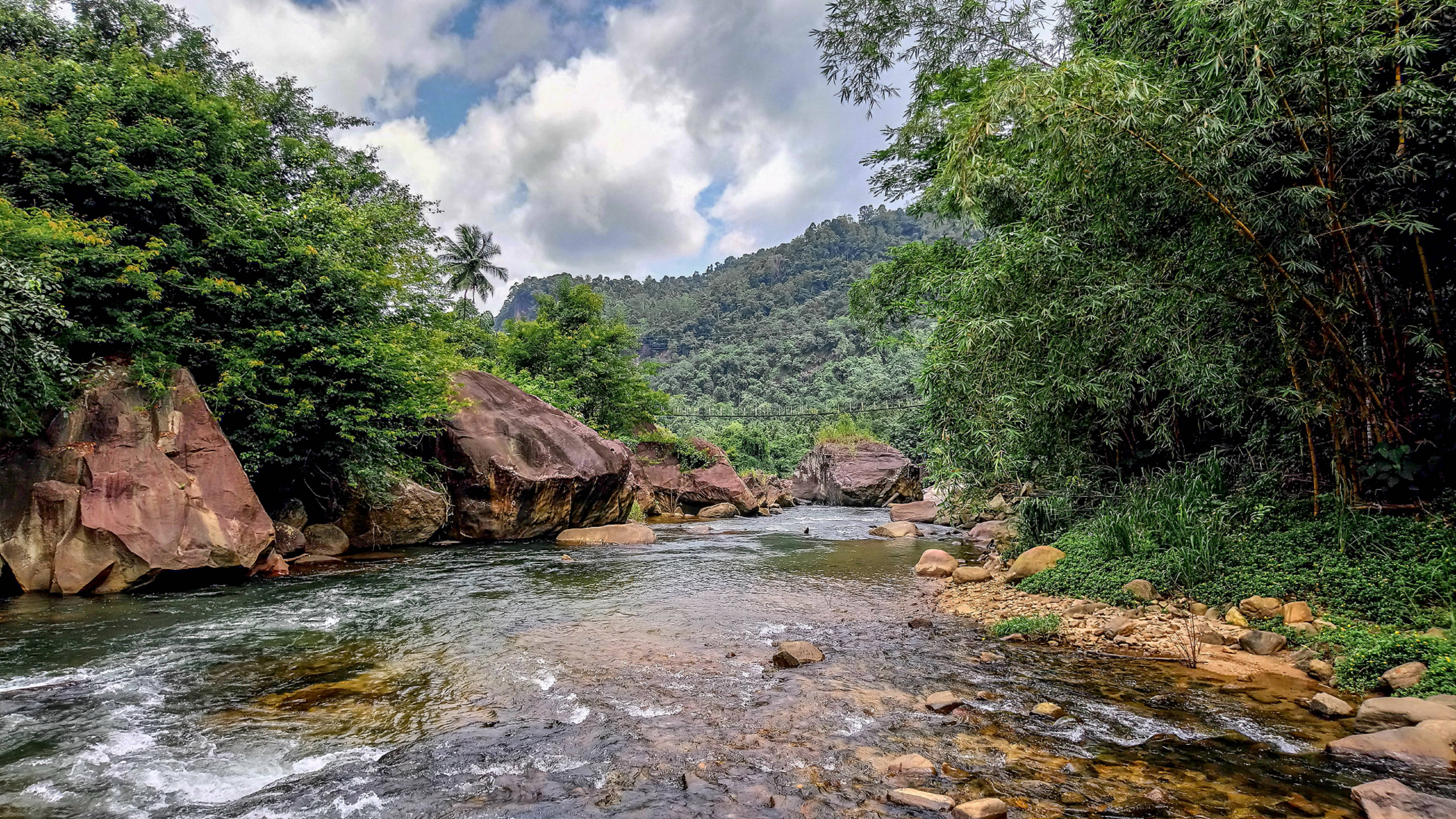 Sri Lanka 桌布 壁紙 — 清澈的河流流過光滑的石頭，穿過鬱鬱蔥蔥的綠色森林，有高聳的竹子和遠處的山丘，天空部分多雲。