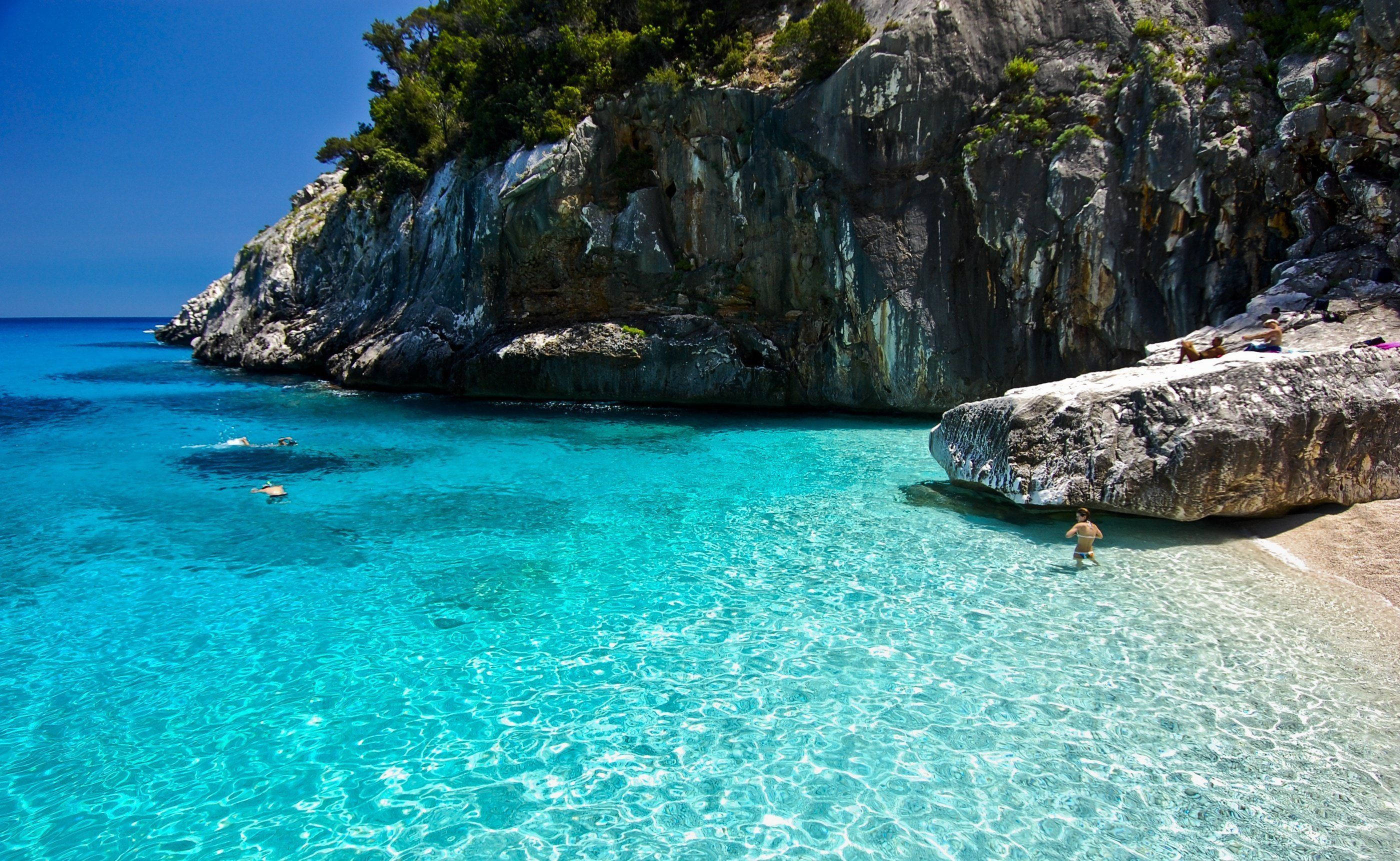 Capri 桌布 壁紙 — 清澈的碧綠海水拍打著僻靜的岩石海灘，天空湛藍，游泳者享受陽光。