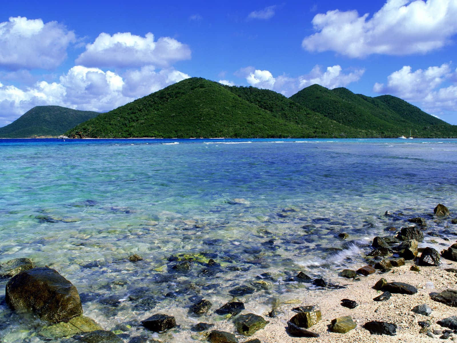 National Geographic Desktop 桌布 壁紙 — 清澈的碧綠海水拍打著岩石海岸，鬱鬱蔥蔥的綠色山丘在明亮的藍天下聳立，點綴著蓬鬆的白雲。