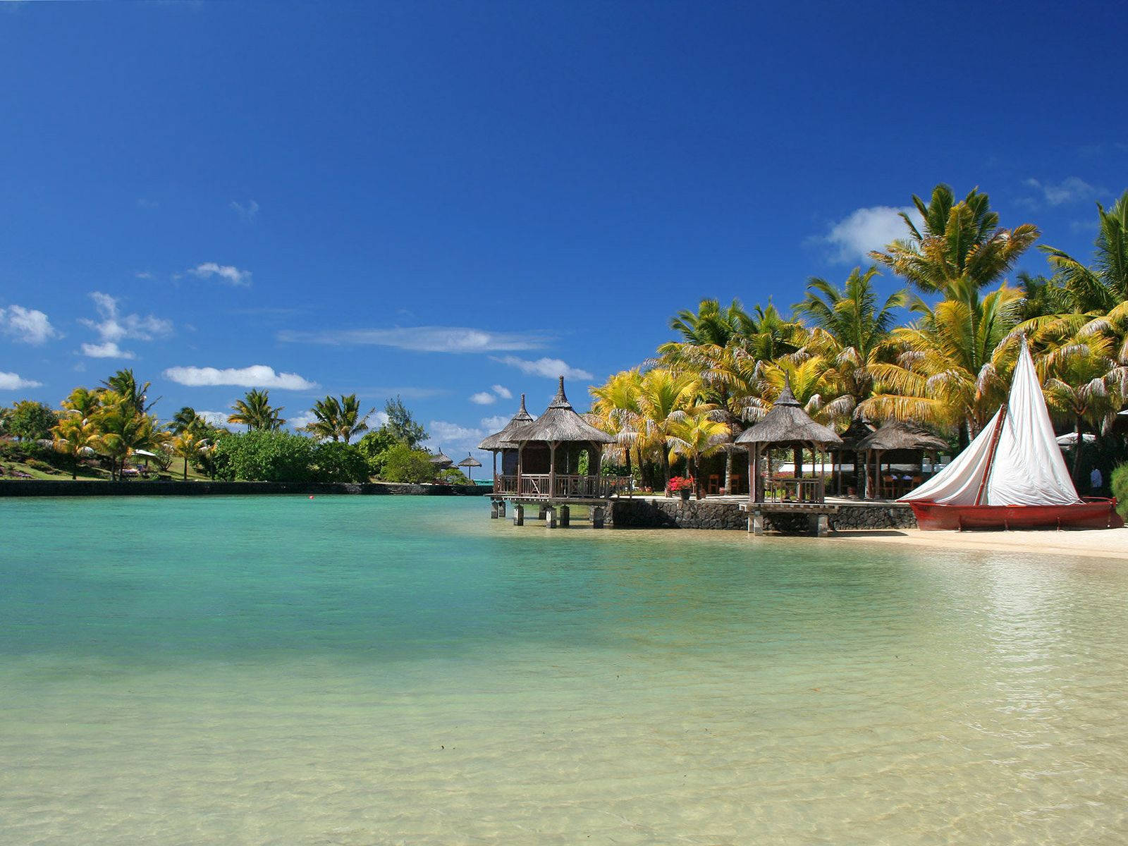 Mauritius 桌布 壁紙 — 清澈的碧綠海水拍打著沙灘，沙灘上棕櫚樹和茅草屋頂小屋林立，頭頂是明亮的藍天。