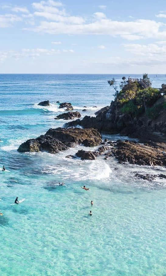Beach Aesthetic 桌布 壁紙 — 清澈的碧綠海水拍打著深色火山岩，衝浪者和游泳者在陽光明媚的海岸天堂中盡情享受。