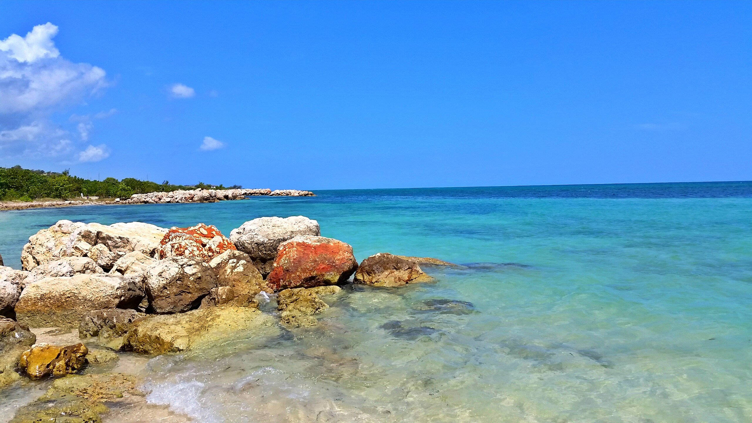 Jamaica 桌布 壁紙 — 清澈的碧綠海水拍打著陽光照射的岩石，天空湛藍，白雲點綴。