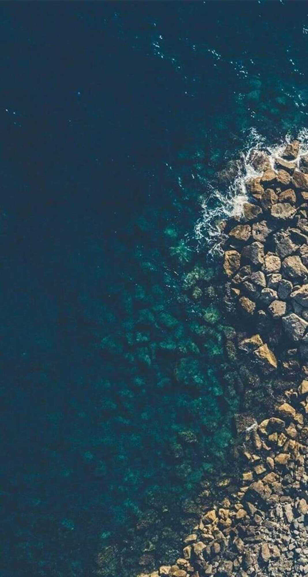 Screen 桌布 壁紙 — 清澈碧綠淺水中，深藍色海浪拍打崎嶇棕色岩石的航拍視圖。