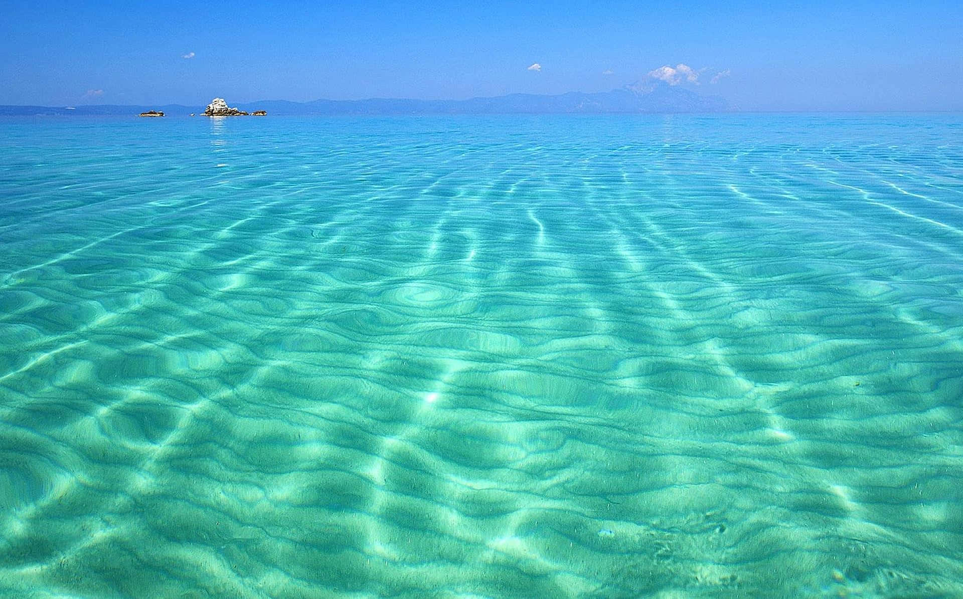 Greek Beach 桌布 壁紙 — 清澈碧綠的海水在明亮的藍天下泛起漣漪，遠處有岩石小島和山脈。